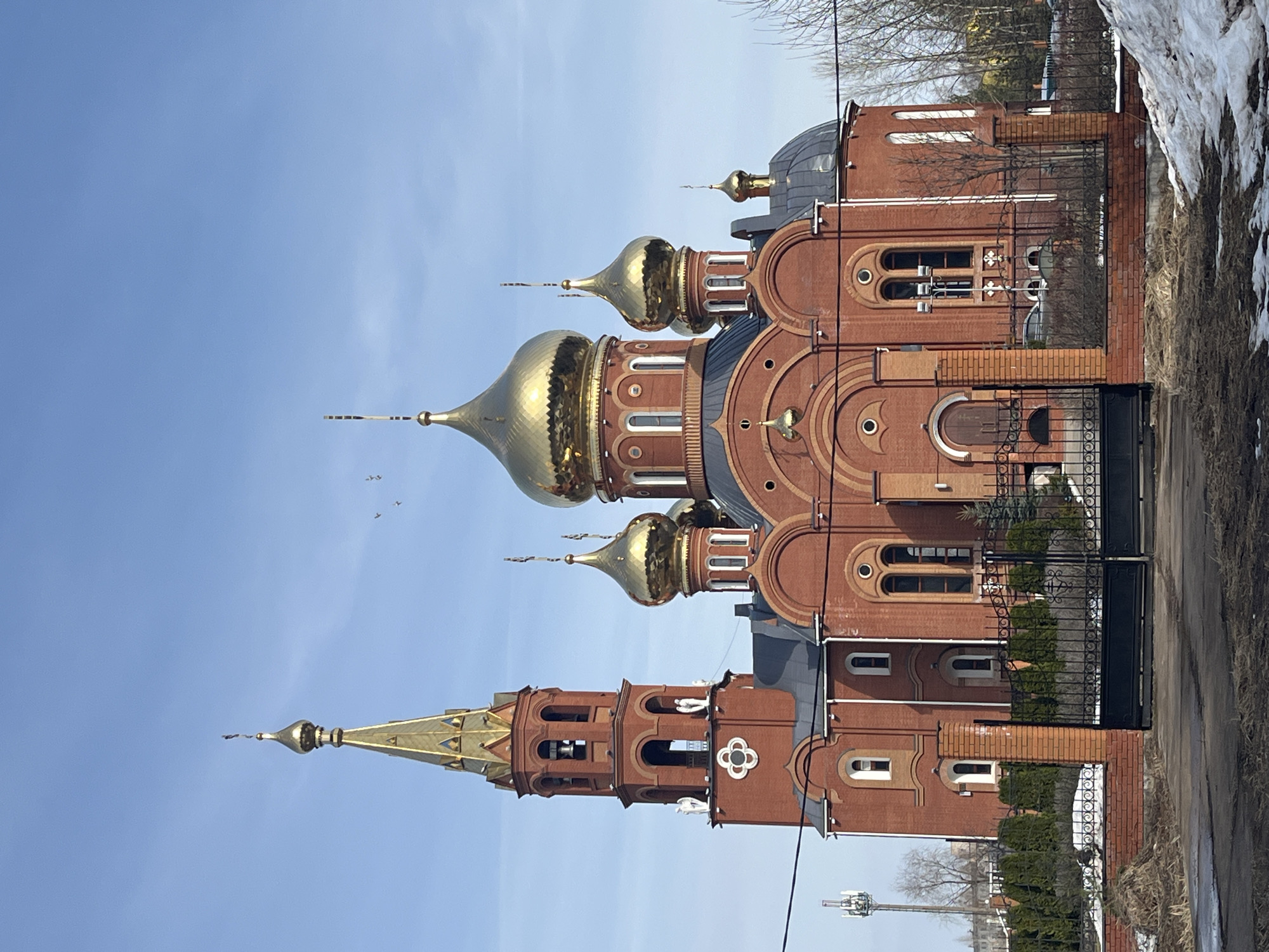 The image shows the Church of the Kazan Icon of the Mother of God in Chaikovsky, Perm Krai, Russia. The church is constructed of red brick and features several golden domes. Some snow patches remain near the church, and a fence surrounds the property. The sky is blue, and some birds are flying in the sky.