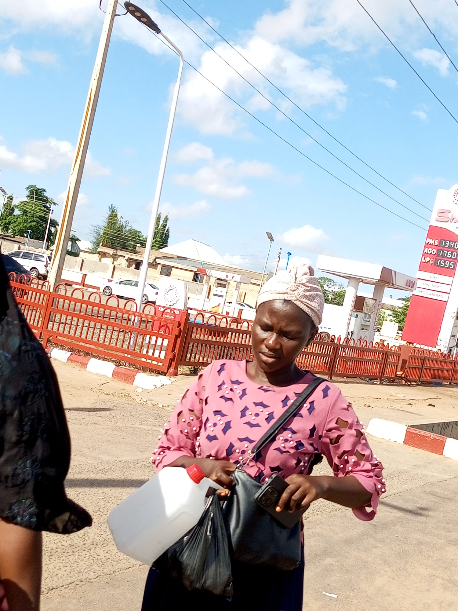 A woman, wearing a pink patterned top and a light-colored headscarf, stands on a paved surface. She is holding a white plastic jerrycan in her right hand and a black bag in her left, which also contains a mobile phone. Behind her, a red metal fence separates the foreground from a street with several parked cars and a gas station with a red and white sign. The sign displays fuel prices, including PMS at 340, AGO at 1360, and LPG at 1595. The sky is blue with scattered clouds. The location is identified as Tumfure, Nigeria.