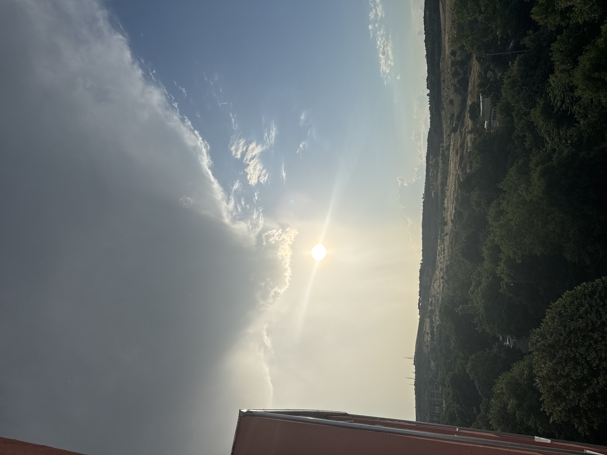 The image depicts an outdoor scene, likely in Burgas, Bulgaria, during daytime. The sky is a prominent feature, with a mix of bright sunlight and clouds. The sun is visible, casting a glare and illuminating the sky with a warm, bright light, suggesting it's either late morning or early afternoon. To the left, a large, dark cloud bank contrasts with the brighter, partly cloudy sky. To the right, a hilly landscape is covered with dense green trees and vegetation. In the distance, some structures and possibly roads are visible amidst the greenery, with power lines also dotting the hillside. The foreground shows the edge of a reddish-brown building with a light-colored roofline, indicating an elevated vantage point for the photograph. The overall atmosphere is bright and natural, with no discernible activity or specific event taking place. There is no visible text in the image.