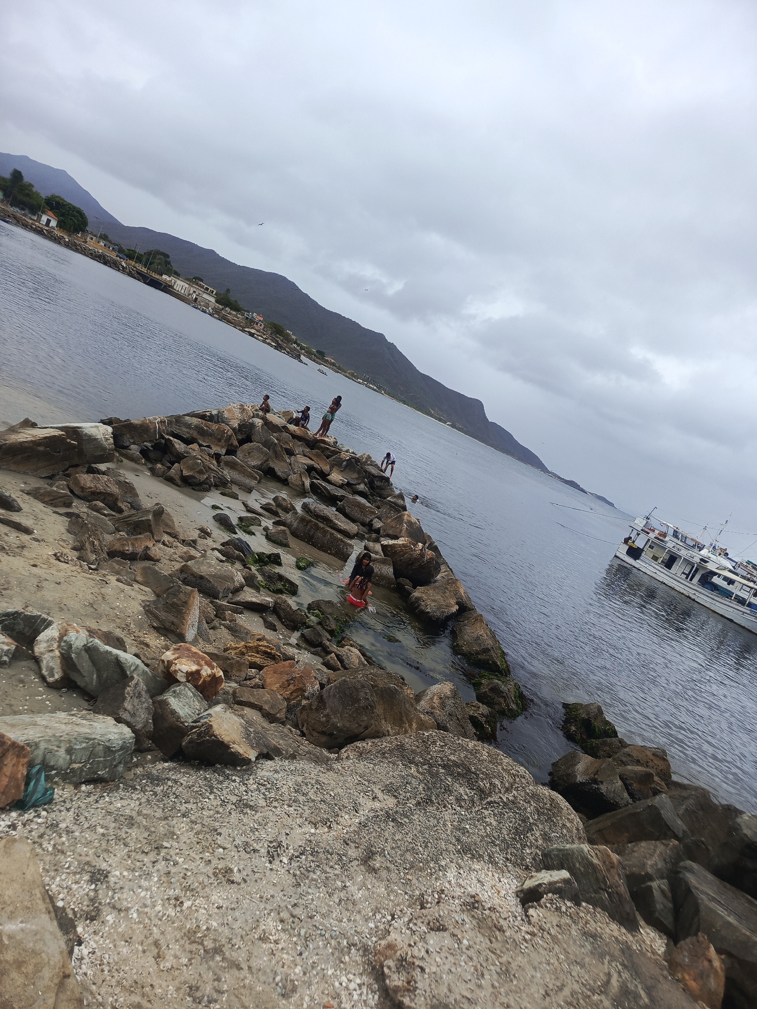 The image depicts a rocky shoreline in Juan Griego, Venezuela, under an overcast sky. The foreground is dominated by large, rough-hewn rocks and sandy areas littered with shells. In the midground, several people, including children, are present on the rocks near the water's edge. Some appear to be wading or playing in the shallow water. A white boat with multiple decks and railings is anchored or moored in the calm, grey water to the right of the frame. The background features a mountainous landscape with some scattered buildings and vegetation, characteristic of the coastal scenery in the area. The overall impression is a casual, outdoor scene by the sea.