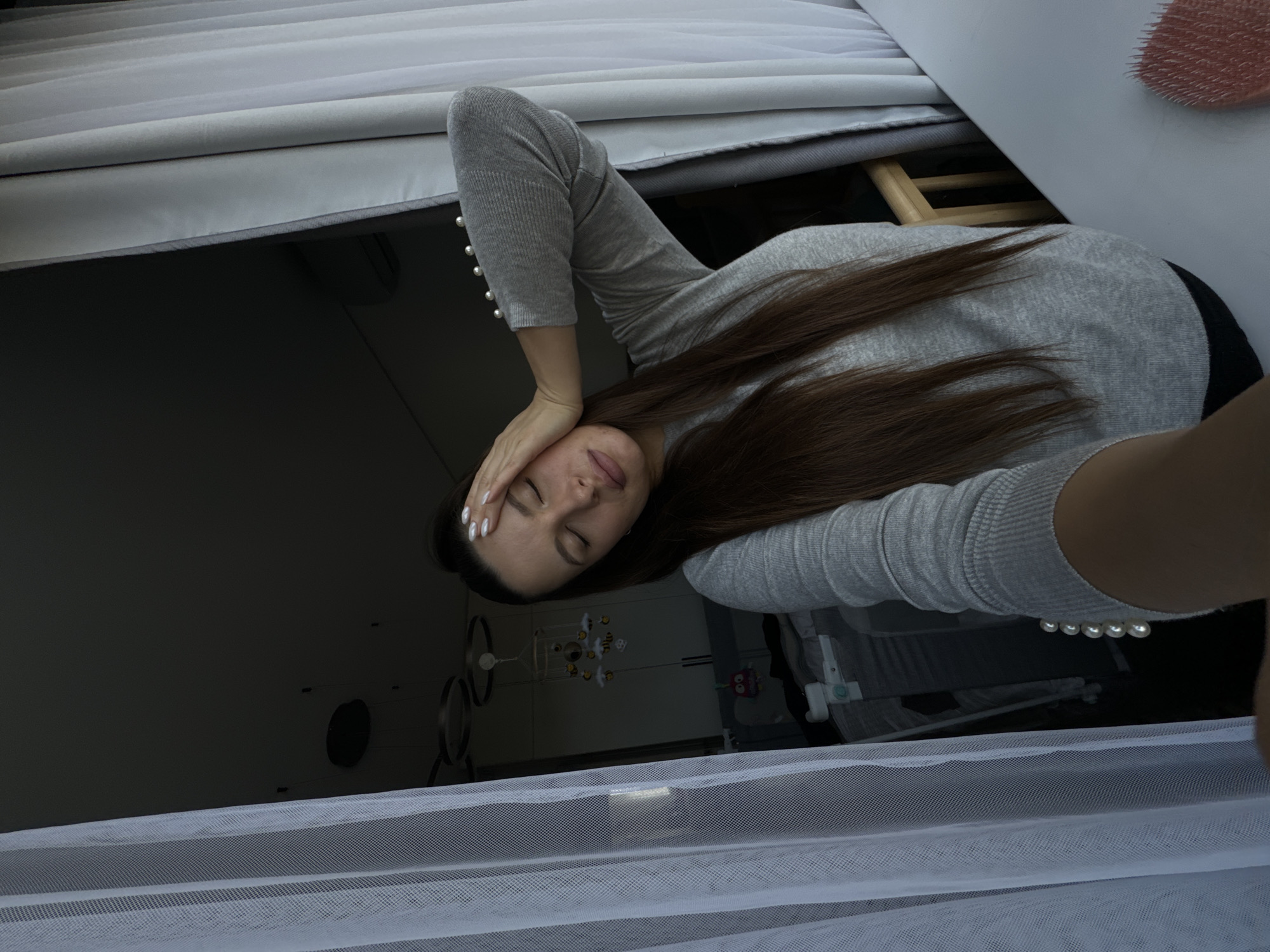 A woman with long brown hair is lying on a grey upholstered bed. She is wearing a grey long-sleeved shirt with pearl details on the sleeves. Her left hand is resting on her forehead. The background is a dark room with a crib and a baby mobile visible through a white mesh fabric. The scene appears to be indoors, possibly a bedroom, in the vicinity of Akulovo (Gp Kubinka), Russia.
