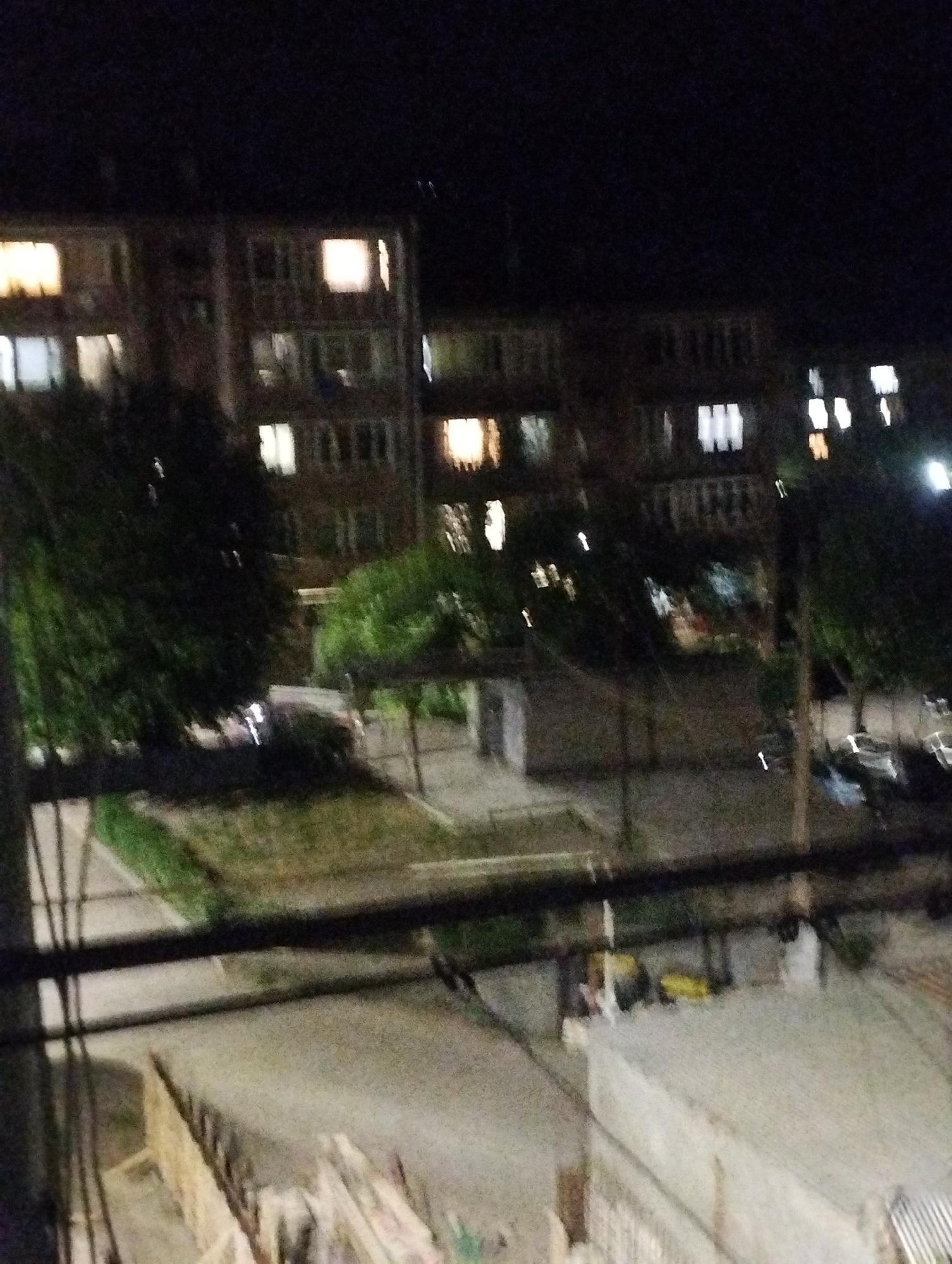 The media file displays a nighttime scene of a residential area in Yeghegnadzor, Armenia, taken from an elevated position, possibly a balcony, as indicated by the prominent railings and cables in the immediate foreground.
The primary subjects are several multi-story apartment buildings, their dark facades punctuated by numerous illuminated windows. The lights within the windows vary in color, ranging from warm yellow and orange to cooler white and blue tones, suggesting different light sources inside the residences. The buildings appear to be typical of urban residential architecture.
Between and in front of the buildings, dense trees and other vegetation are visible, some obscuring parts of the structures. Ground areas consist of paved surfaces, possibly roads or paths, and grassy patches. In the midground, what appear to be parked cars or distant vehicles with their lights on are faintly discernible. Near the bottom of the frame, indistinct objects, possibly construction materials like wooden planks, are present on the ground.
The scene depicts a quiet, ordinary night in a residential neighborhood. No specific activity or event is taking place; the illuminated windows simply indicate the presence of people inside the buildings. The time of day is clearly night, with a dark, cloudless sky. The image has a grainy and somewhat blurry quality, consistent with low-light photography. No visible text can be identified in the image.