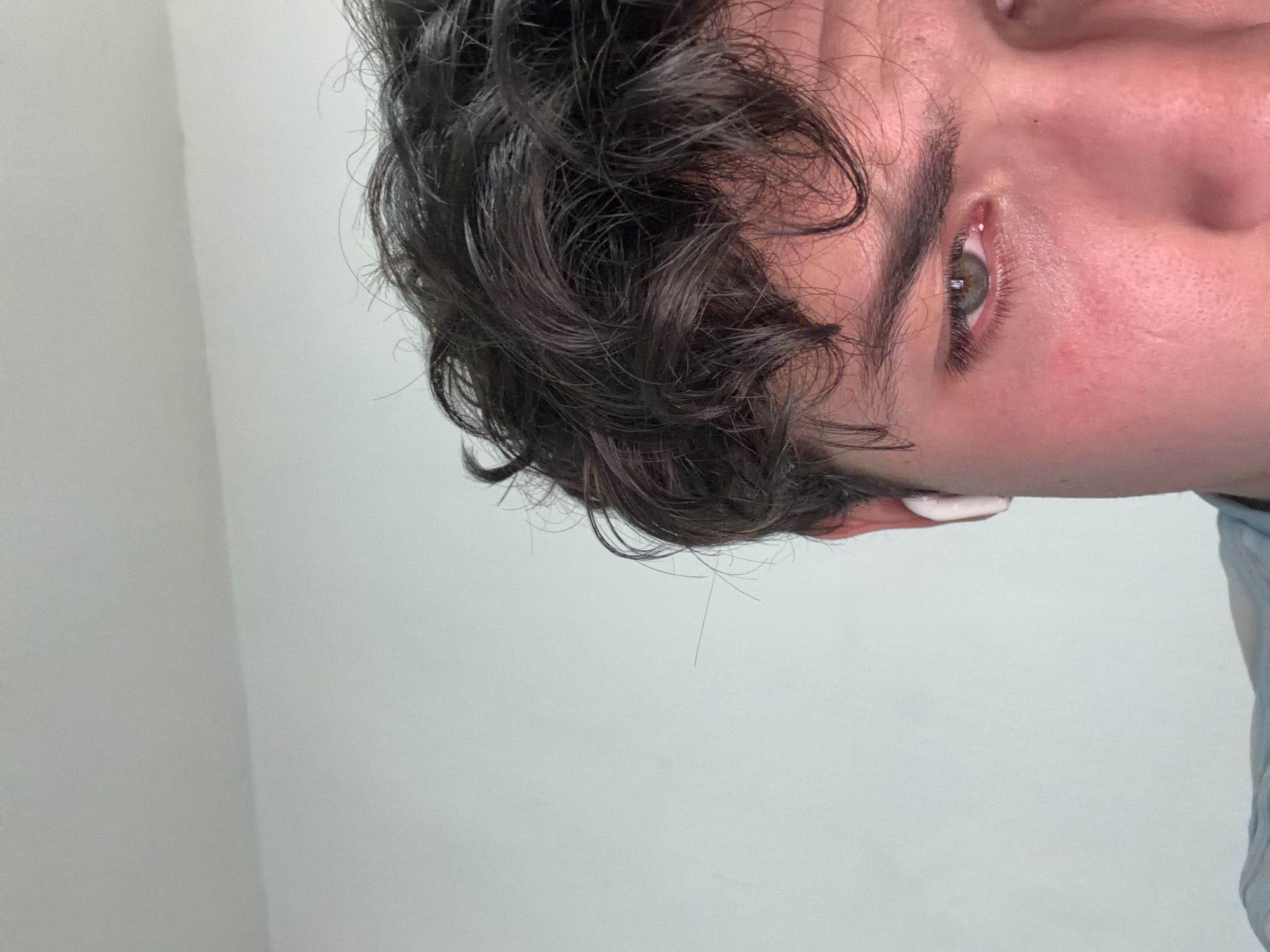 A close-up, rotated image focuses on the right side of a person's face. The individual has dark, curly hair, light skin with slight redness around the eye, and an eyebrow with a somewhat unkempt appearance. The visible eye is hazel in color. A small amount of white substance is visible on the ear, potentially cream or lotion. The person wears a light blue shirt. The background is a plain, light gray wall. There is no discernible information to indicate the location is San Andrés Cholula, Mexico.