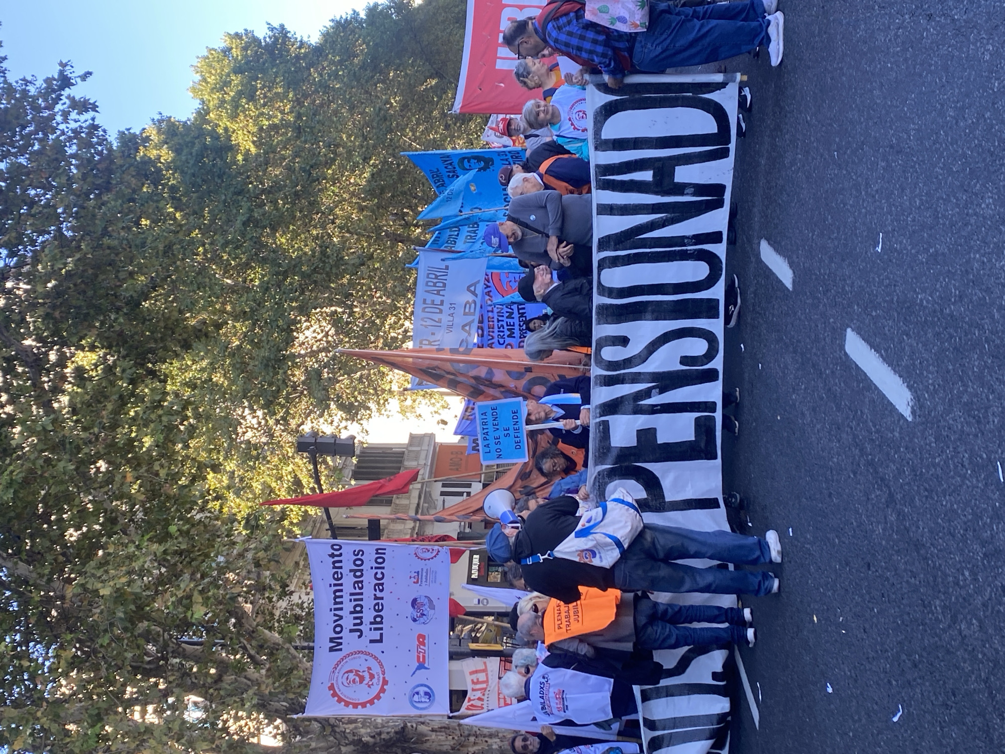 A group of people are protesting in a street, holding banners and flags, likely in Buenos Aires, Argentina, based on the signage language. The banners say "PENSIONAD," "Movimiento Jubilados Liberacion," "LA PATRIA NO SE VENDE SE DEFIENDE," and "R-12 DE ABRIL ABA VILLA 31". The protesters are mostly elderly. The environment includes trees and buildings. The street is marked with white paint lines.
