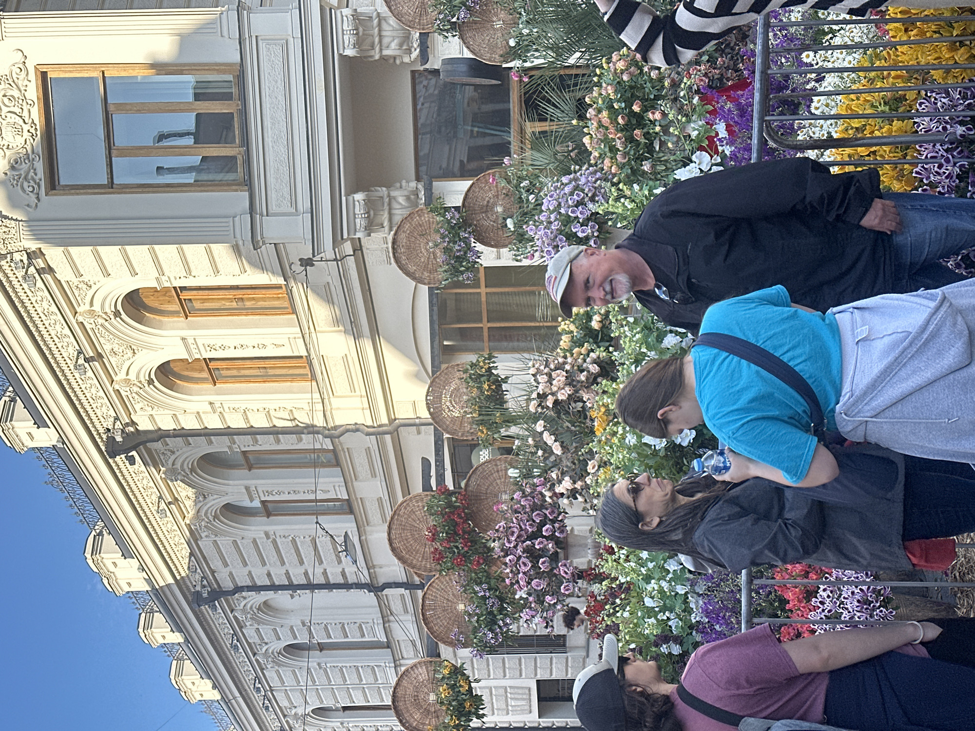 A street scene displays a group of four individuals amidst decorative floral arrangements, set against the backdrop of an ornate building facade under a clear blue sky. The individuals appear to be tourists or locals observing and interacting with the floral display. They are dressed in casual clothing suitable for warm weather, indicative of a possible outdoor activity or sightseeing.
The building features classical architectural details, including arched windows, decorative moldings, and a facade embellished with intricate designs. Wicker baskets filled with colorful flowers line the building's exterior, adding to the aesthetic appeal.
The floral arrangements and the ornate building suggest a possible tourist destination or a well-maintained urban area. The exact location within the city of Unknown, Unknown, is indistinguishable based on the image alone.