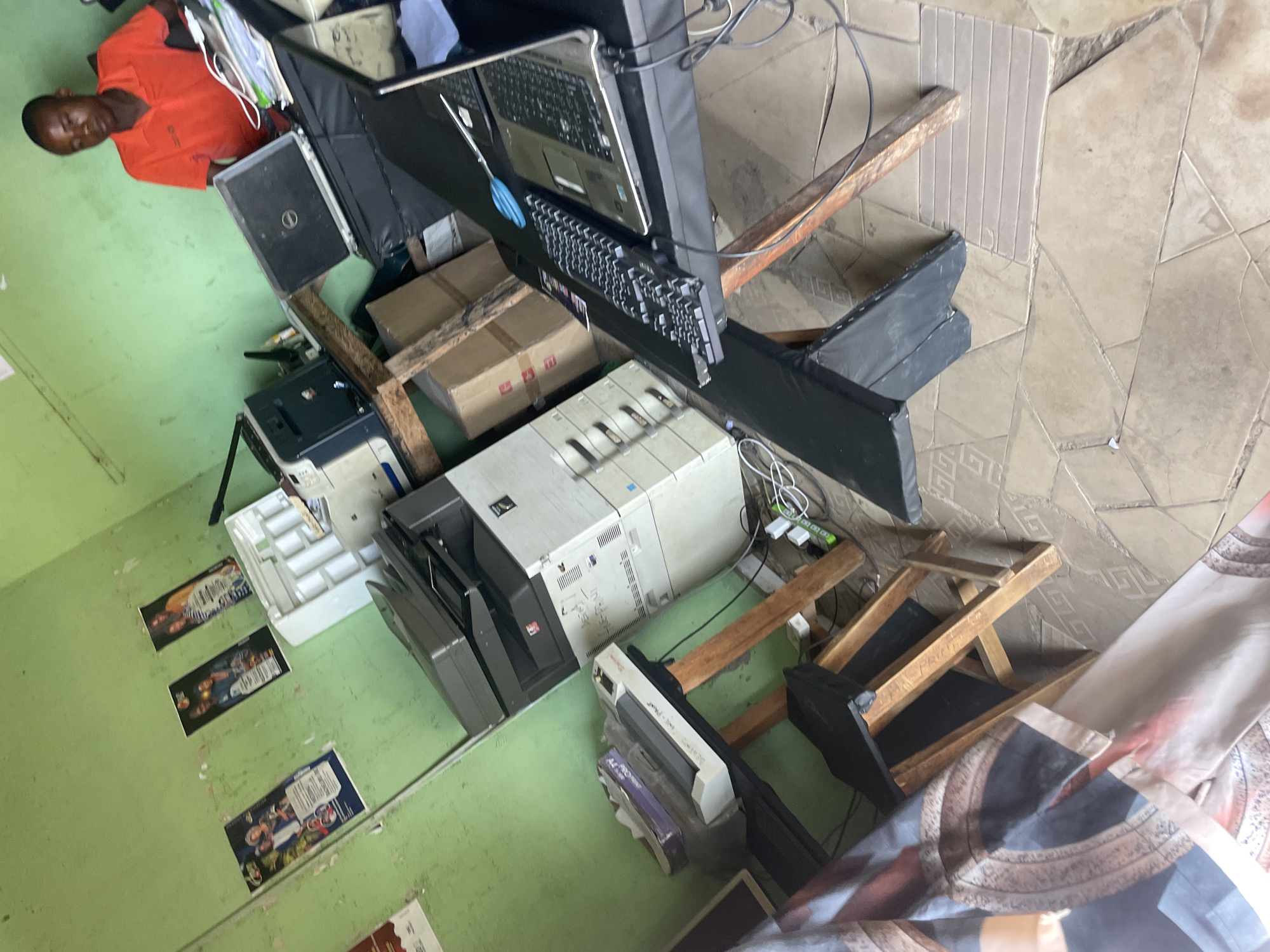 The image depicts an indoor workspace in Mararaba, Nigeria. A man wearing an orange shirt is partially visible in the upper left. Multiple printers and copiers are present, stacked atop wooden structures. Boxes, computer equipment (laptops, keyboard), and cables are also visible. The floor is green and has posters attached to the wall. The flooring material in the top right transitions to tiled flooring. A patterned cloth is in the foreground.