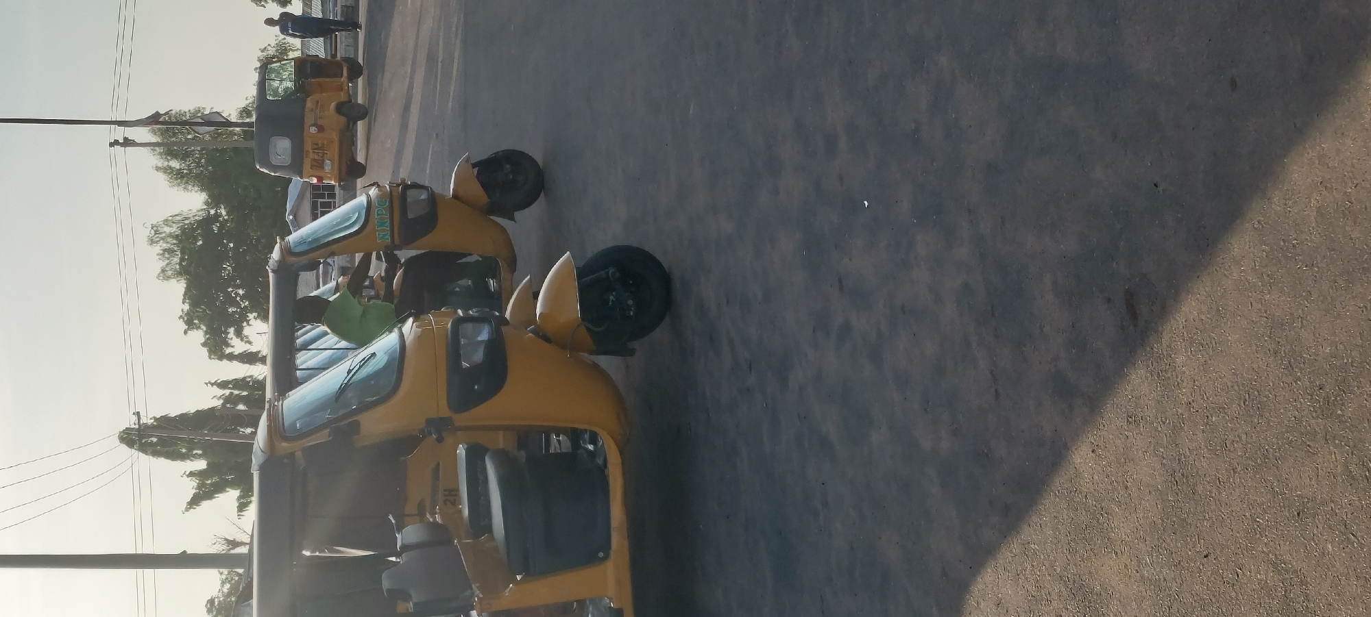 Two yellow three-wheeled auto-rickshaws are parked on an asphalt road in Jalingo, Nigeria. The auto-rickshaws have black roofs and black seats. One auto-rickshaw is in the foreground, on the left, and is partially visible. The second auto-rickshaw is behind it, on the right, and is fully visible. A man in a green shirt is sitting in the driver's seat of the second auto-rickshaw. In the background, a third yellow auto-rickshaw is seen further down the road, along with a man standing next to it. Trees and power lines are visible in the background under a clear sky. The lighting suggests it is daytime, likely late afternoon due to the long shadows.