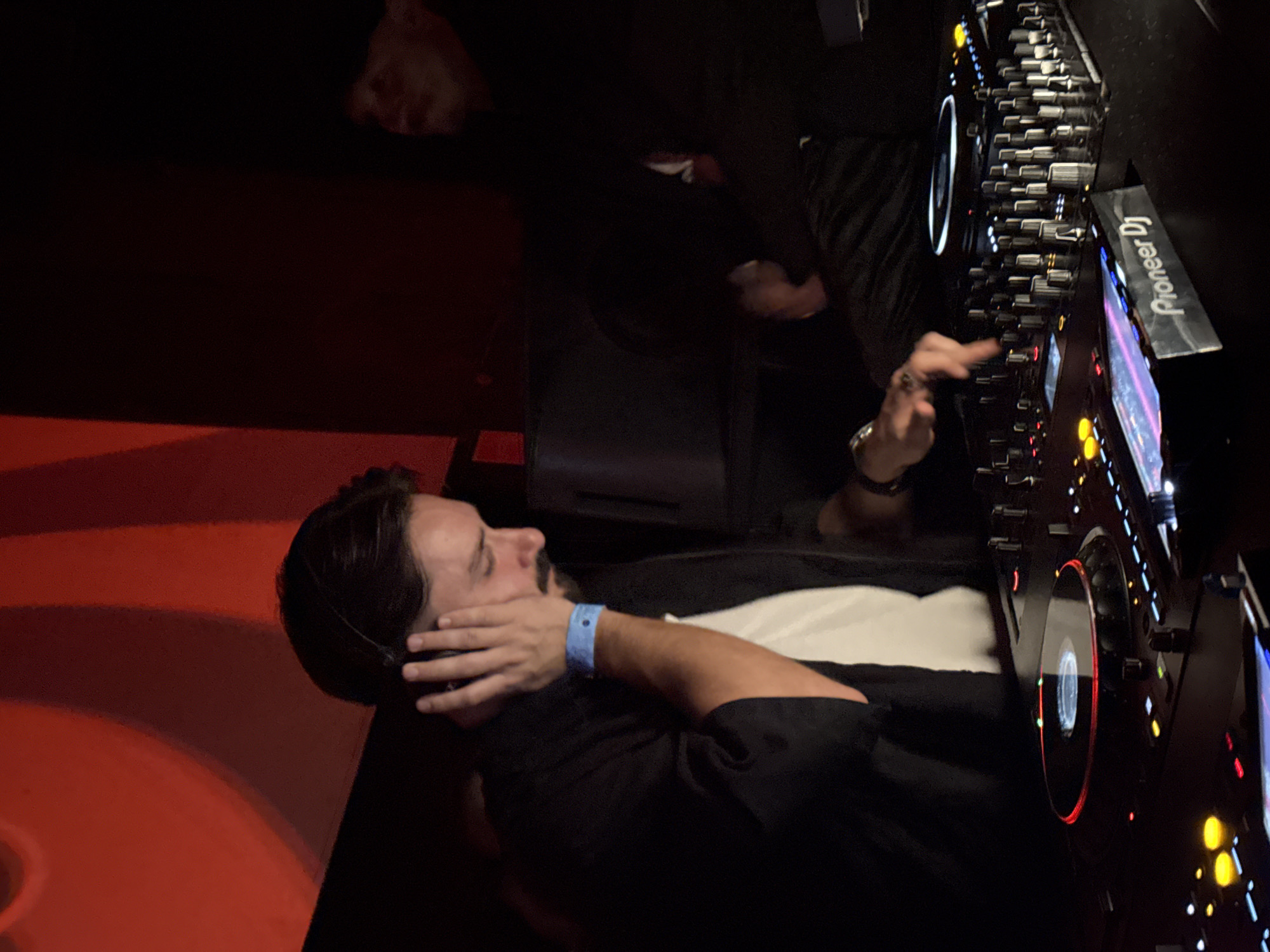 A person, likely a disc jockey (DJ), is positioned at a Pioneer DJ console. They are wearing a black shirt over a white undershirt and have a blue wristband on their left wrist. The console is black and has illuminated buttons and screens. The environment has red lighting and appears to be a nightclub in Barcelona, Spain.