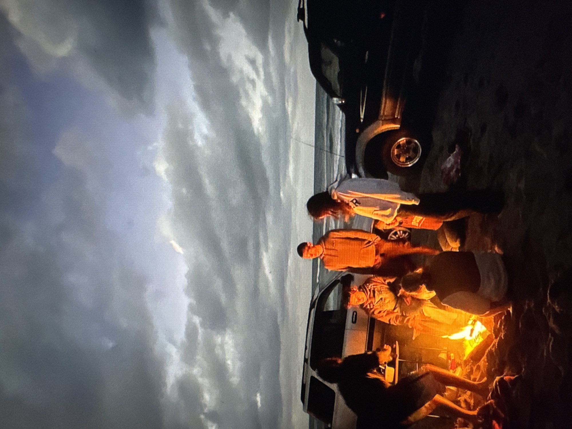 The image depicts a nighttime scene on a beach, with a group of people gathered around a bonfire. Two SUVs are parked nearby, one white and one black. The sky is cloudy, with hints of blue and grey. The fire is bright and casts a warm glow on the people and the surrounding sand.
The people in the image appear to be young adults, dressed in casual clothing. Some are sitting, while others are standing. They seem to be engaged in conversation and enjoying the warmth of the fire.
The location is identified as the city of Tallinn, Estonia. However, the image itself does not contain any visual cues that specifically confirm this location. The scene could be any beach with a similar environment. The overall atmosphere is one of relaxation and camaraderie.