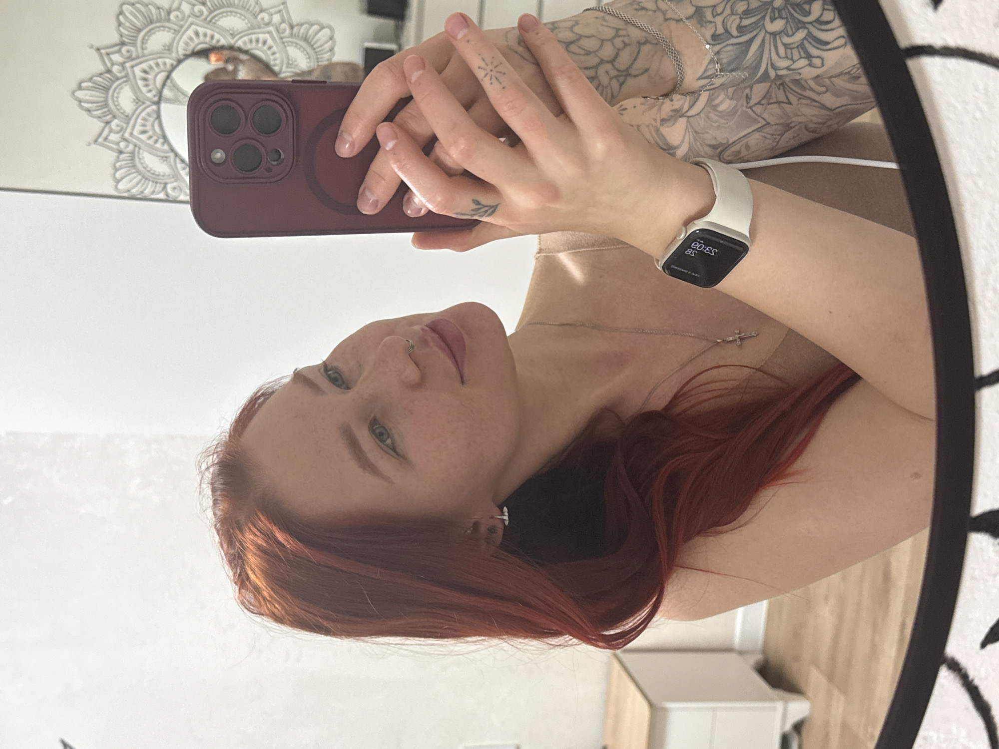 A medium close-up mirror selfie captures a young woman with reddish-brown hair and freckles, looking directly into the reflection of her smartphone camera. Her expression is neutral. She is wearing a light-colored top and has several pieces of jewelry, including a silver chain necklace with a cross pendant and a small nose hoop piercing.
Her hands hold a dark red/burgundy smartphone, likely an iPhone with a triple camera system and a visible MagSafe ring. Extensive grey-scale tattoos, depicting intricate floral or mandala-like patterns, cover her left arm and shoulder. Smaller, delicate tattoos are visible on her fingers: a star-like design on her right middle finger and a leaf or branch on her left ring finger. On her right wrist, she wears a white Apple Watch, its screen visible though mirrored, showing "00:ES" (likely 00:53 or 5:00) and "E`00:8S" (likely 08:00) along with reversed text "SNNENOONVO," which is possibly a notification.
The reflected background behind the woman includes a white wall and a decorative, grey mandala-like pattern. The bottom right of the reflection shows part of a light wood floor and a white piece of furniture, possibly a dresser or cabinet. The edge of the mirror's frame is visible on the right side of the image. The scene is indoors, likely in a room in Rostov-on-Don, Russia.