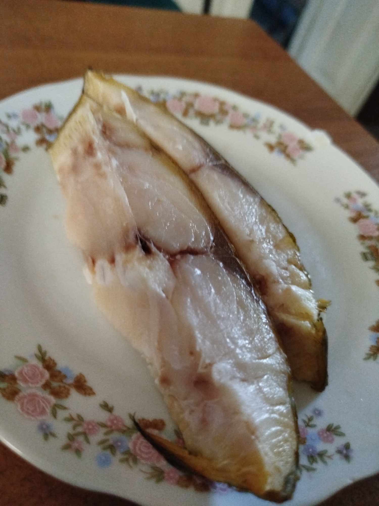 The image shows two pieces of cooked fish on a white plate with a floral pattern. The fish is light brown and appears flaky. The plate is on a wooden surface. The scene is likely from a kitchen or dining area. There is no indication of the location in Nova Znamianka, Ukraine.