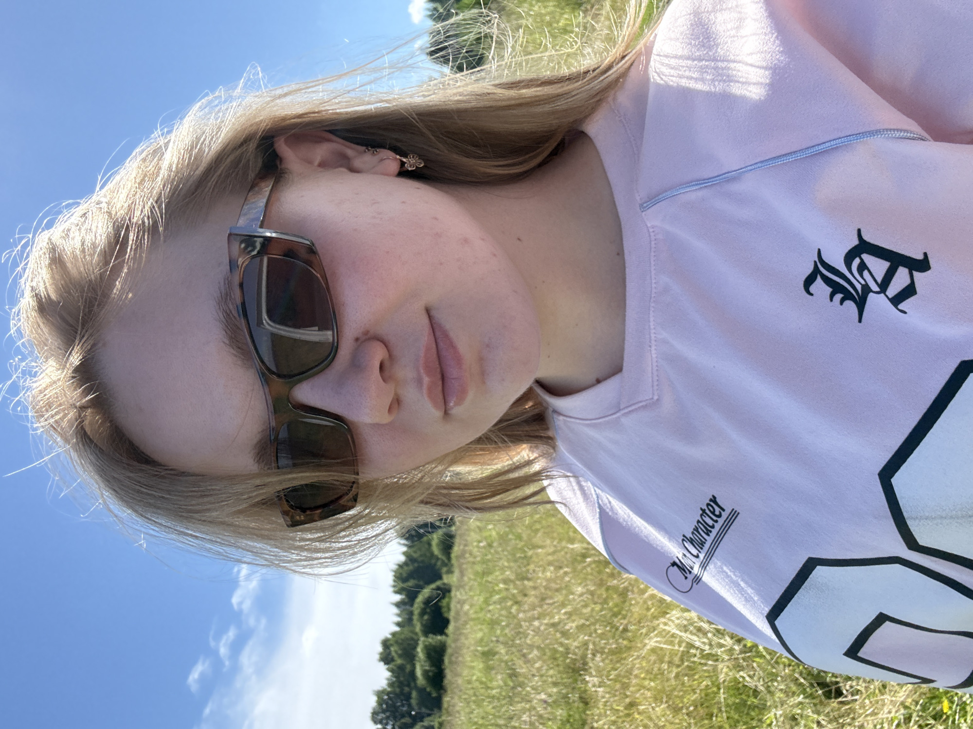 A young woman with blonde hair and sunglasses is taking a selfie in a grassy field. She is wearing a pink sweatshirt. The background is a clear blue sky with some clouds, and a line of trees in the distance. The location is the city of Kryukovo, Russia.