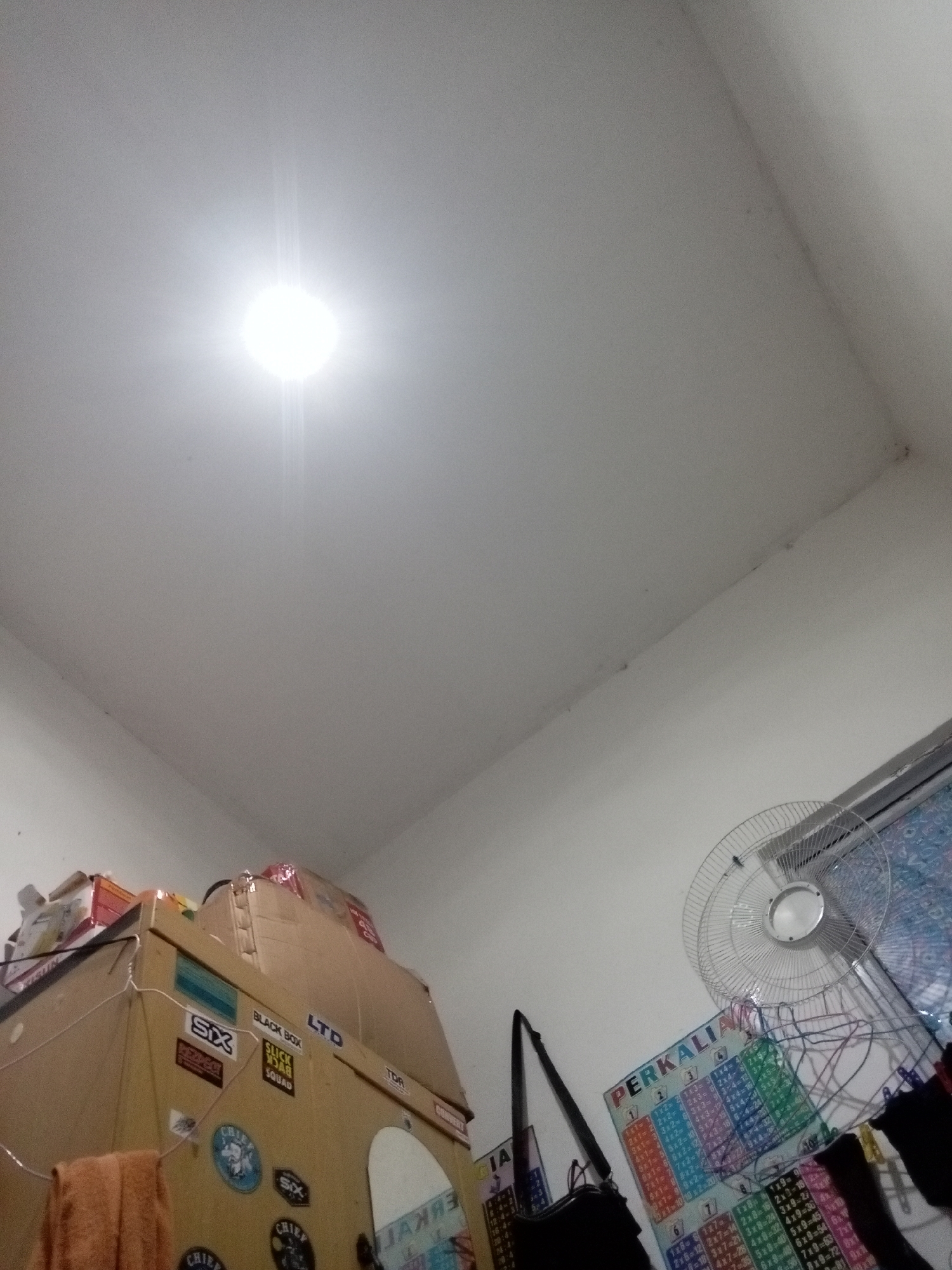This image depicts an indoor scene, likely a room in Batam, Indonesia, given the presence of Indonesian language on a multiplication chart. The perspective is from below, looking upwards towards a white ceiling with a bright light fixture.
In the lower left portion of the image, a tall, light brown cardboard wardrobe or storage unit is visible, adorned with various stickers, including "SIX", "BLACK BOX", "LTD", and circular emblems with text. An orange towel is draped over the front of this unit.
To the right of the wardrobe, a white oscillating fan is positioned on what appears to be a window ledge or shelf. Behind the fan, a window with a patterned blind is partially visible. Below the fan, a colorful chart with Indonesian text "PERKALIAN" (multiplication) is displayed on the wall. The chart features multiplication tables.
In the foreground, to the left of the fan, a black shoulder bag is hanging. The walls appear to be painted white. The overall environment suggests a domestic or potentially a small office space. No individuals are present in the frame.