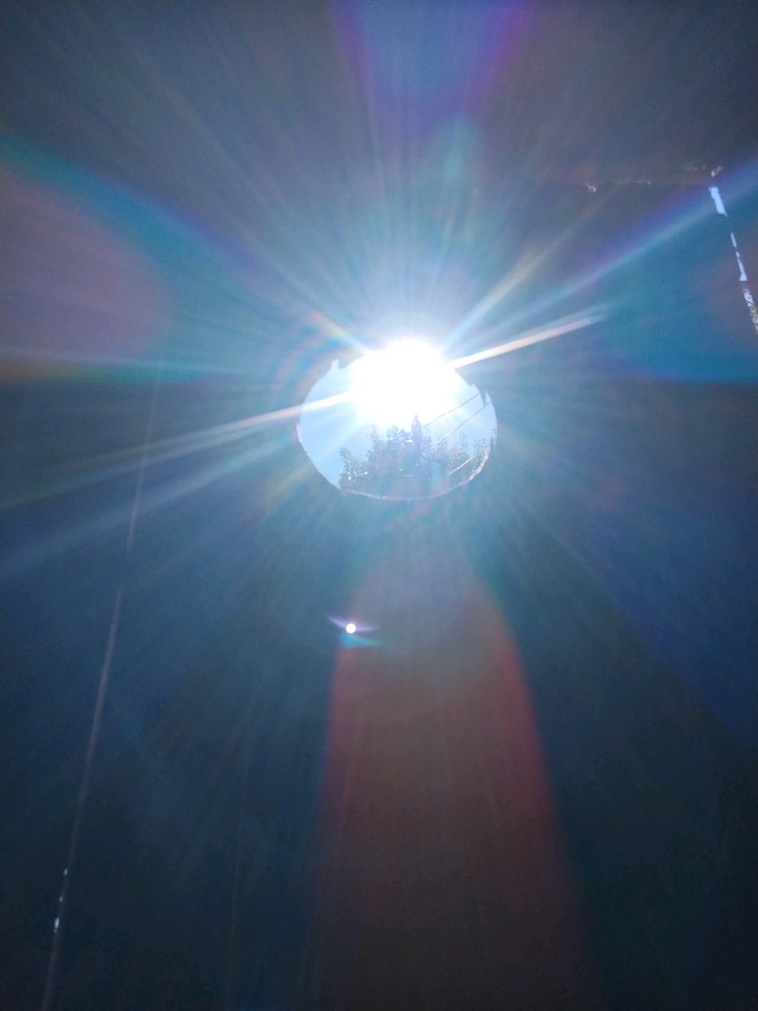 A close-up, low-angle shot captures the sun's intense glare, creating numerous light flares and a lens flare effect. The sun itself is partially obscured by a circular opening, through which a faint reflection of trees and sky is visible. The background is predominantly dark blue and black, with streaks of blue and purple light emanating from the sun. A smaller, white light source appears lower in the frame, also surrounded by a reddish-brown glow. The scene is devoid of any people or specific urban landmarks that would place it in Hoholeve, Ukraine. The composition suggests an outdoor setting, possibly looking up through a skylight or aperture.