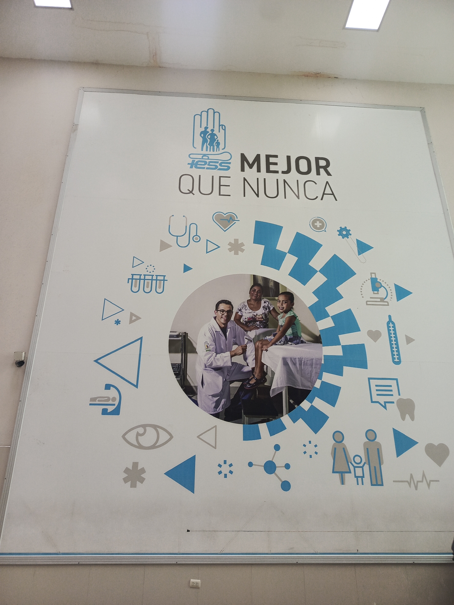 This image shows a large mural on a wall, likely in Guayaquil, Ecuador, given the presence of the IESS logo. The mural features a medical theme with various icons related to healthcare, such as a stethoscope, heart rate monitor, microscope, and a family silhouette. In the center of the mural, a doctor in a white lab coat is examining a young girl sitting on a medical examination table. A woman is also present, seated behind the girl. The background of the mural is white, with abstract blue and grey shapes and medical graphics. The overall impression is one of a health awareness campaign or an advertisement for a medical institution. The wall appears to be indoors, possibly in a clinic or hospital waiting area. Above the mural, a section of the ceiling shows signs of water damage.