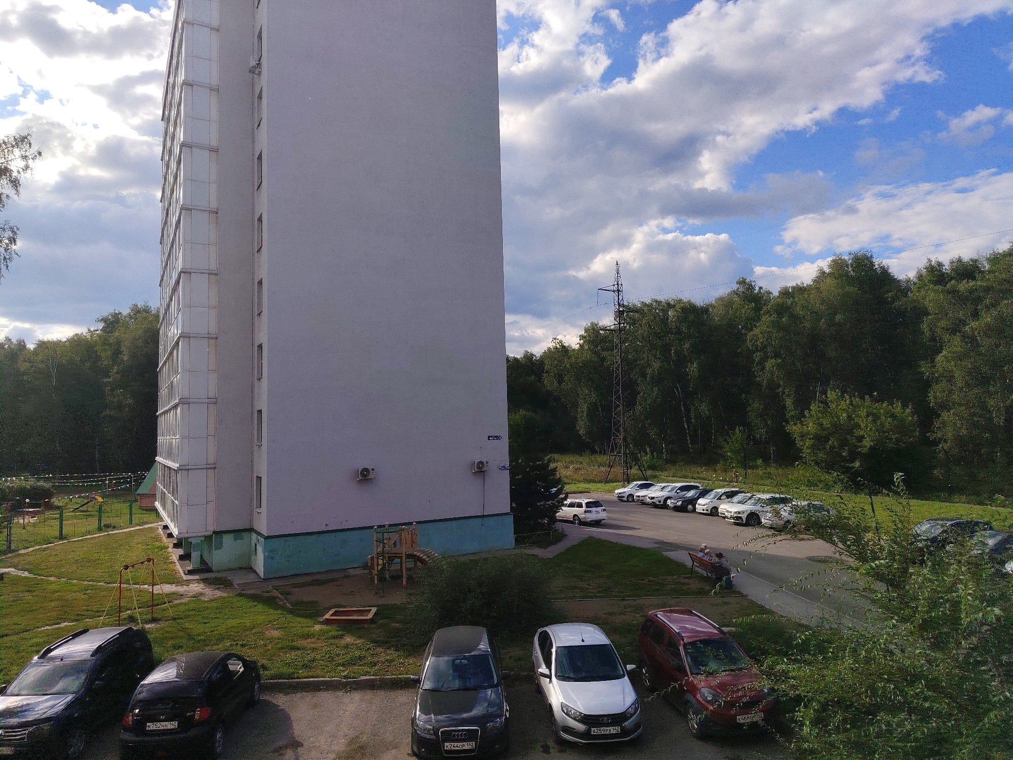 This is a medium-angle, high-contrast outdoor shot of a residential building and its surroundings, taken from a higher vantage point, possibly an apartment window. The prominent, multi-story building occupies the left half of the frame, featuring a stark white facade and numerous balconies with glass railings. To the right of the building, a dense forest forms the background, meeting a bright blue sky punctuated by scattered white clouds. A power line tower and wires are visible amongst the trees. In the foreground, a parking lot is filled with various cars, including a black SUV, a black sedan, a silver sedan, and a red SUV. A park area with a playground is visible in the middle ground, between the building and the trees. The overall scene depicts a typical urban residential environment with elements of nature and recreation. Based on the license plates of cars in the image (e.g., K752HH 142, K244OP 142, A259PB 142, K468EM 142), the location is Kemerovo, Russia.