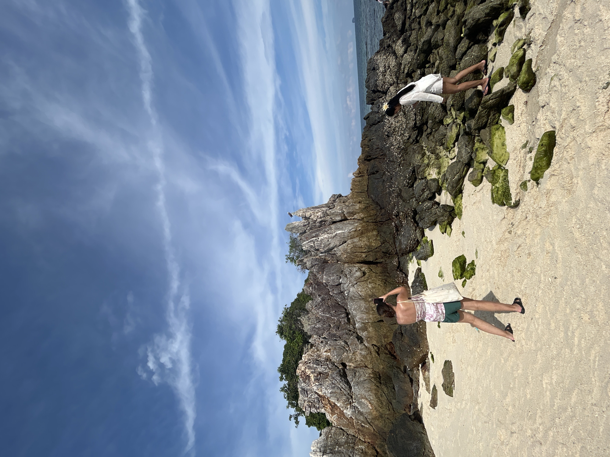 The image captures a serene coastal scene at Phru Nai, Thailand, under a clear blue sky dotted with wispy white clouds. In the foreground, a sandy beach transitions into a rocky shoreline covered in patches of green moss. Two individuals are present: a woman in the mid-ground, facing away from the camera, is taking a photograph of the rocky landscape with her phone. She wears a patterned top, green shorts, and flip-flops, and carries a tote bag. To her right, a second woman, also facing away, is walking on the rocks. She is dressed in a white outfit and has a yellow flower in her hair. In the background, a large, rugged rock formation rises from the sea, topped with lush green foliage. The ocean stretches to the horizon, where a distant island is faintly visible.
