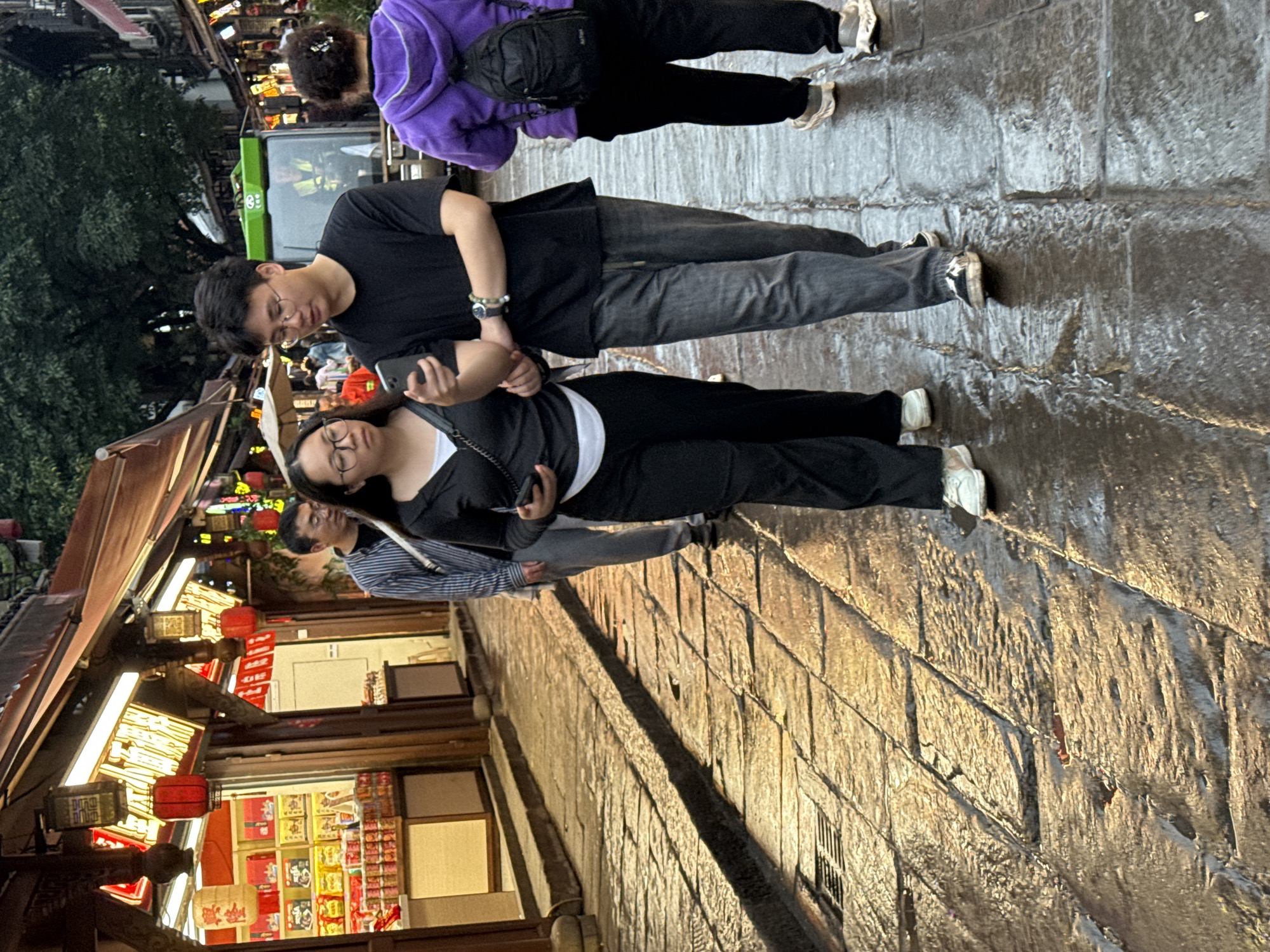 The image shows an outdoor scene on a stone-paved street in Shapingba District, China. Several individuals are walking on the street. One person is wearing a purple jacket and black pants. A couple is standing together, both looking at cellphones. The man wears a black shirt and gray pants, while the woman is in a black top and black pants. Another individual in a striped shirt is partially visible behind them. Shops line one side of the street, adorned with hanging red lanterns and lit displays of goods. A portion of a green bus is also visible in the background. The ground appears wet, suggesting a recent rain or cleaning.
