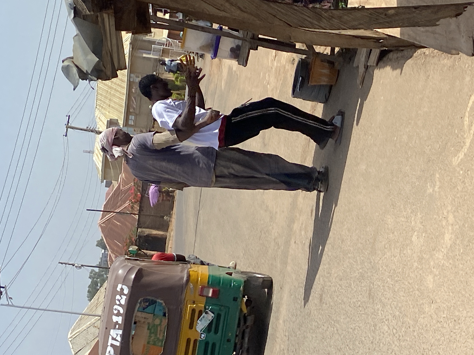 The image captures an outdoor street scene in what appears to be a bustling area of Jos, Nigeria, during bright daylight. Two men are featured prominently in the foreground, seemingly interacting near a makeshift stall.
The man on the left, possibly older, wears a light-colored headwrap, a grey short-sleeved shirt, and dark trousers. He is partially obscured by the second man and appears to be facing him. The man on the right, younger, is dressed in a white t-shirt with a red trim at the bottom, black track pants with two white stripes down the sides, and light-colored sandals. His hands are raised and brought together in front of him, suggesting an action like clapping or holding a small item. This interaction takes place next to a simple wooden and corrugated metal structure, likely a roadside stall, under which plastic containers are visible, one holding what looks like small, round fruits. A dark bucket is also nearby.
In the mid-ground to the left, a three-wheeled vehicle, commonly known as a "keke Napep" (tricycle taxi), is parked. It has a brown canopy and a yellow and green body. The text "PTA-1923" is clearly visible in white lettering on its side.
The background shows residential buildings under a clear, bright sky, indicative of sunny weather. Utility poles and overhead power lines crisscross the upper left portion of the image. Strong, defined shadows are cast on the dusty ground, suggesting it is midday or early afternoon. The overall scene depicts a typical daily street life in an urban or semi-urban setting.