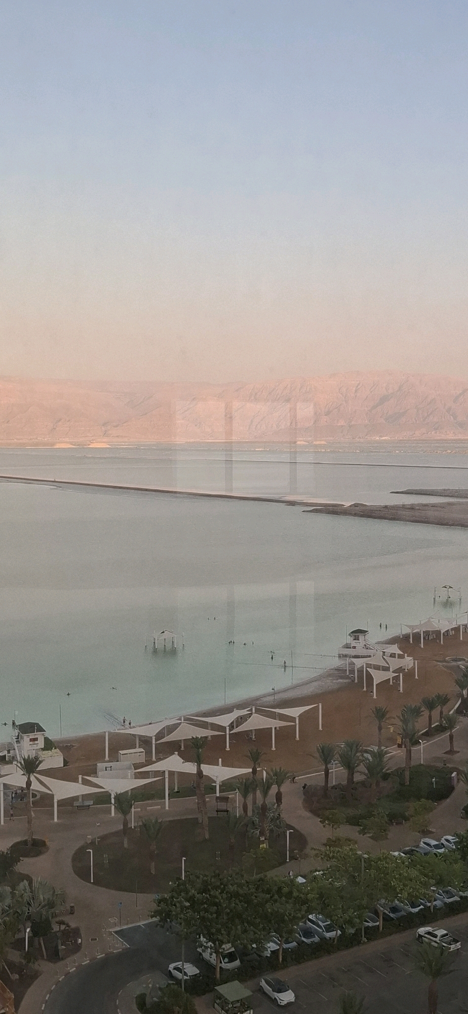 The image shows a coastal resort area with a beach, the Dead Sea, and a parking lot. The beach is lined with white umbrellas and palm trees. The Dead Sea is a large body of water with a calm surface. In the distance, mountains can be seen. A parking lot filled with cars is located near the beach, and there are also some people walking and swimming in the sea. This scene is likely located in the Tamar Regional Council, Israel, given the presence of the Dead Sea.