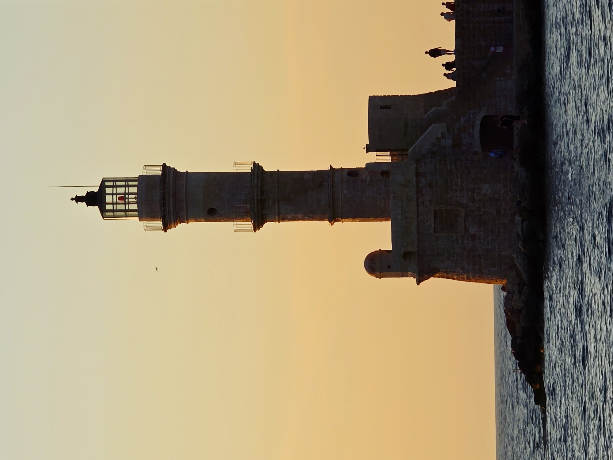 The image depicts the Venetian Lighthouse of Chania, Greece, at sunset. The lighthouse, a tall, cylindrical stone structure with a lantern room at the top, stands on a rocky pier jutting into the sea. The sky is a gradient of warm orange and yellow hues, indicative of a sunset. The sea is dark blue and choppy.
Several people are visible on the pier, silhouetted against the sky. Some are standing, looking out at the sea, while others appear to be walking or gathered in groups. One small bird is flying in the upper left portion of the sky. The lighthouse itself is illuminated by the setting sun, casting a warm glow on its stone facade. The base of the lighthouse has a more fortress-like structure with small turrets and arched doorways. The overall scene is serene and picturesque, capturing the beauty of the ancient landmark during golden hour.