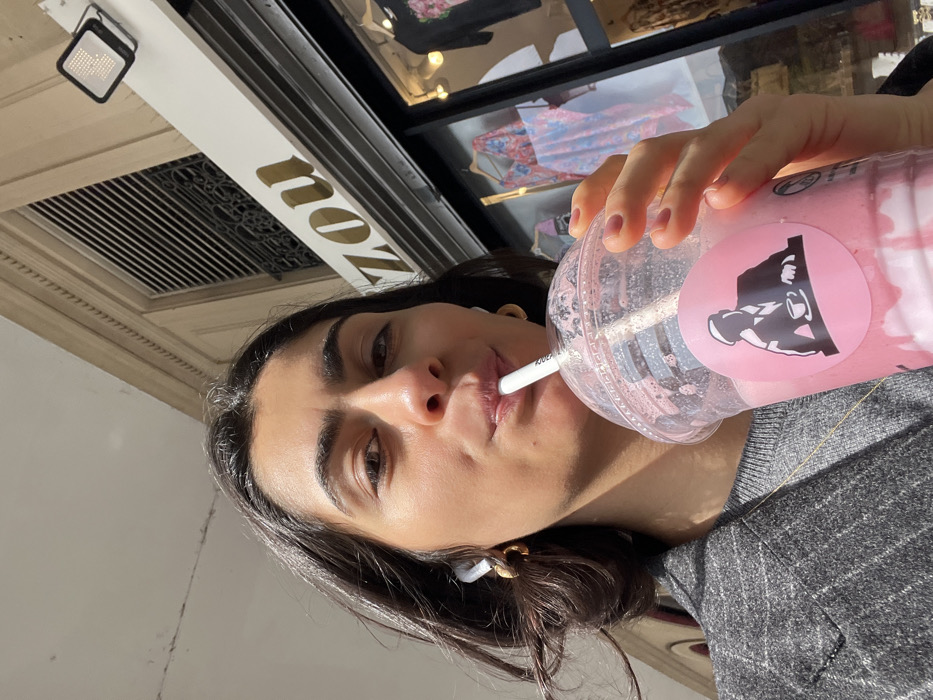 A female subject is depicted in a low-angle close-up, looking directly at the camera while actively drinking from a clear plastic cup through a white straw. She has dark hair, wears small gold hoop earrings, and white wireless earbuds. Her right hand, with pink-painted fingernails, holds the cup, which contains a pink layered beverage with white liquid at the bottom and features a circular pink sticker with a black silhouette of a person in a cap sitting at a table with a cup. She is dressed in a grey striped blazer over a grey sweater, with a thin gold chain visible around her neck. In the background, a storefront displays clothing through a large window. Above the display, a gold-lettered sign partially reads "Zou." An outdoor floodlight is affixed above the sign. The building exhibits classic Parisian Haussmannian architectural elements, including stone facades, mouldings, and a grated window. This scene is located in Paris, France, specifically in front of the "ZOU" store at 8 Rue Montorgueil, 75001.
