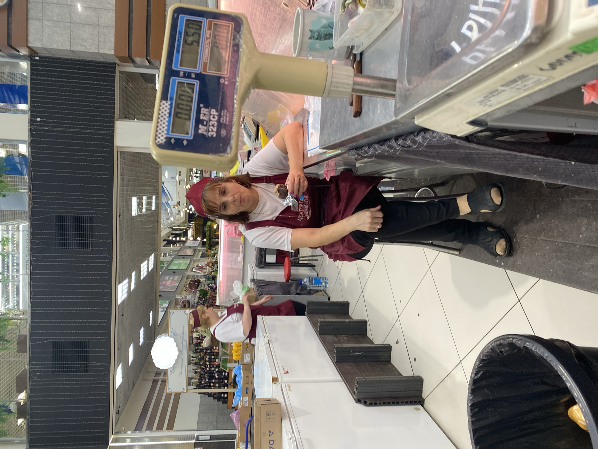 The image shows a retail setting, likely a grocery store or market, in Novaya Usman', Russia. A woman wearing a red hat and a burgundy apron over a white shirt stands behind a counter. She is holding a chocolate bar in her right hand.
In front of her is a weighing scale with a blue digital display. The display shows "5.50" in the upper section and "0.000" in the lower section. Text on the scale reads "M-ER 323CP". A sign above the scale shows Russian text.
The background reveals aisles stocked with products, including shelves of bottled drinks and what appear to be packaged goods. Fluorescent lights illuminate the store. A large white refrigerator is visible to the right of the woman. A black trash can is also in the foreground. The floor is tiled in a diamond pattern.
The image appears to be taken during daylight hours, likely within a store. The woman's expression is neutral. The overall scene depicts a typical retail environment where a product is being weighed or prepared for sale.
