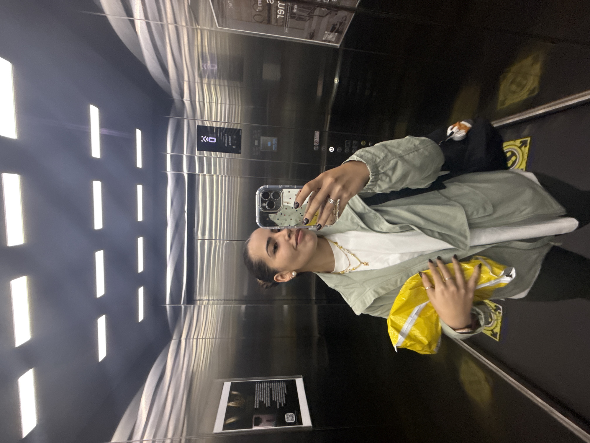 The image depicts a young woman taking a selfie in an elevator. The elevator has metallic walls and ceiling with linear lighting. The woman is holding a smartphone with a clear case. She is wearing a light green jacket over a white shirt. She holds a large yellow shopping bag. The location is an elevator, but there is no indication that it is in San Bernardino Tlaxcalancingo, Mexico.