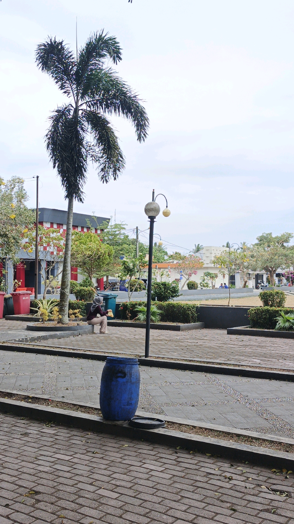 In the city of Desa Karangbenda, Indonesia, an outdoor public space is depicted under an overcast sky. In the foreground, a blue plastic barrel with a black lid lies on its side next to a concrete curb. A paved walkway made of reddish-brown bricks leads away from the viewer, transitioning into a patterned stone walkway. A woman wearing a headscarf and a maroon top and beige pants is crouched down near a palm tree, holding a yellow object in her hands. She is positioned on a raised planter bed filled with green bushes. To her left are two large red trash bins. Behind the woman, a building with a red and white checkered facade and a dark roof is visible, partially obscured by trees. A lamppost with two globe-shaped lights stands to the right of the woman. The background shows more trees, low hedges, and other buildings, suggesting a mixed urban and green environment.
