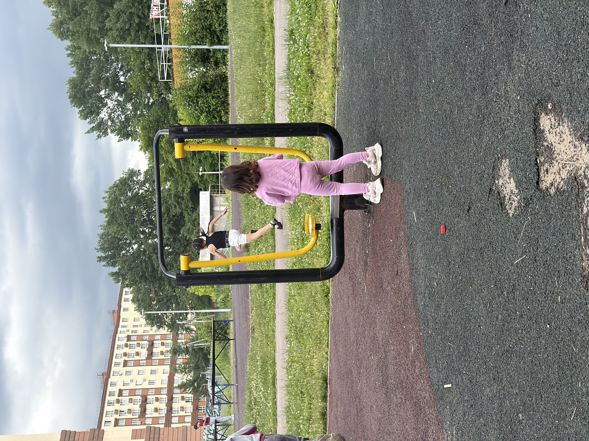 The image depicts an outdoor playground scene in Lopatino, Russia. In the foreground, a young girl wearing a pink tracksuit and sneakers stands facing away from the camera, observing another child on an exercise machine. The exercise machine is a yellow and black structure with footrests, designed for leg exercises. The second child, positioned on the machine, is wearing a black tank top and white shorts and appears to be in motion.
The playground surface is a mix of dark grey and reddish-brown rubberized material, with patches of wear and tear visible. Scattered on the ground are small debris items, including a small red object.
In the midground, a paved path runs horizontally, bordered by green grass with scattered white clover flowers. Beyond the path, more exercise equipment and a long, dark picnic table are visible.
The background features a large, multi-story residential building with a beige and brown facade, characterized by numerous windows and balconies. Dense green trees form a backdrop to the building and extend across the upper part of the image. The sky is overcast with a cloudy, grey atmosphere, suggesting a possibly cool or rainy day. A street lamp is visible near the building. A person wearing a red shirt and blue jeans is standing near the picnic table, looking towards the exercise equipment.