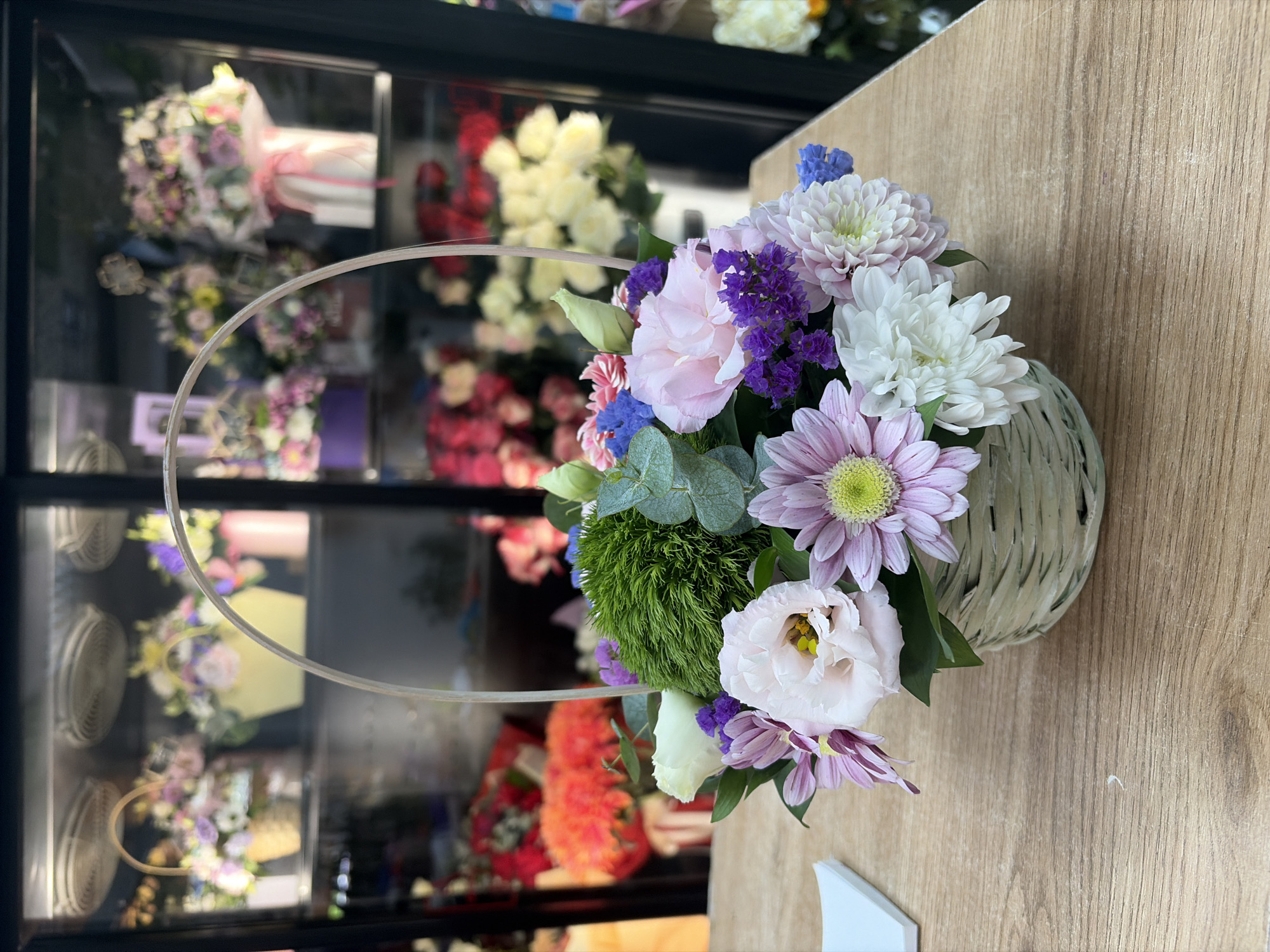 A close-up, eye-level shot displays a light green woven basket filled with a variety of flowers, resting on a light brown wooden surface. The basket's handle, made of clear plastic, arches over the arrangement. In the foreground, the bouquet features large pink and white roses, pink daisies with yellow centers, a spiky green plant, purple statice, and blue aster flowers. The background, visible through a glass display case, shows more floral arrangements in baskets and vases, suggesting the location is a flower shop. The blurred background suggests the image was taken in Budva, Montenegro, likely within a commercial establishment. There are no people visible in the scene.