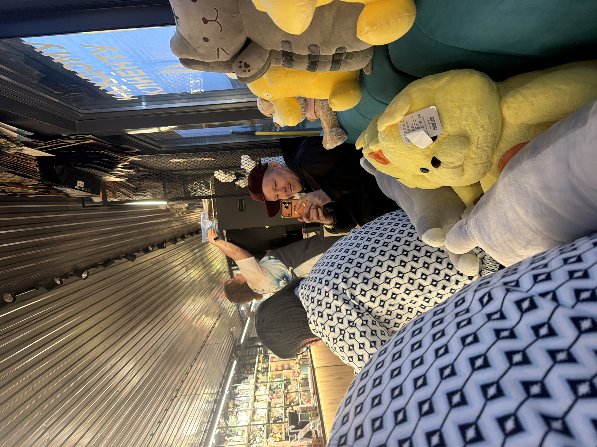 A medium indoor shot from Chaikovsky, Russia, shows a man wearing a maroon cap and black hoodie taking a photo with an orange phone. The man is surrounded by various plush toys including a gray cat, a yellow bear, and other soft objects. He is sitting on a patterned black and white cushion. In the background, shelves filled with merchandise and other people are visible. The ceiling features spotlights, and a sign above suggests it is a commercial establishment.
