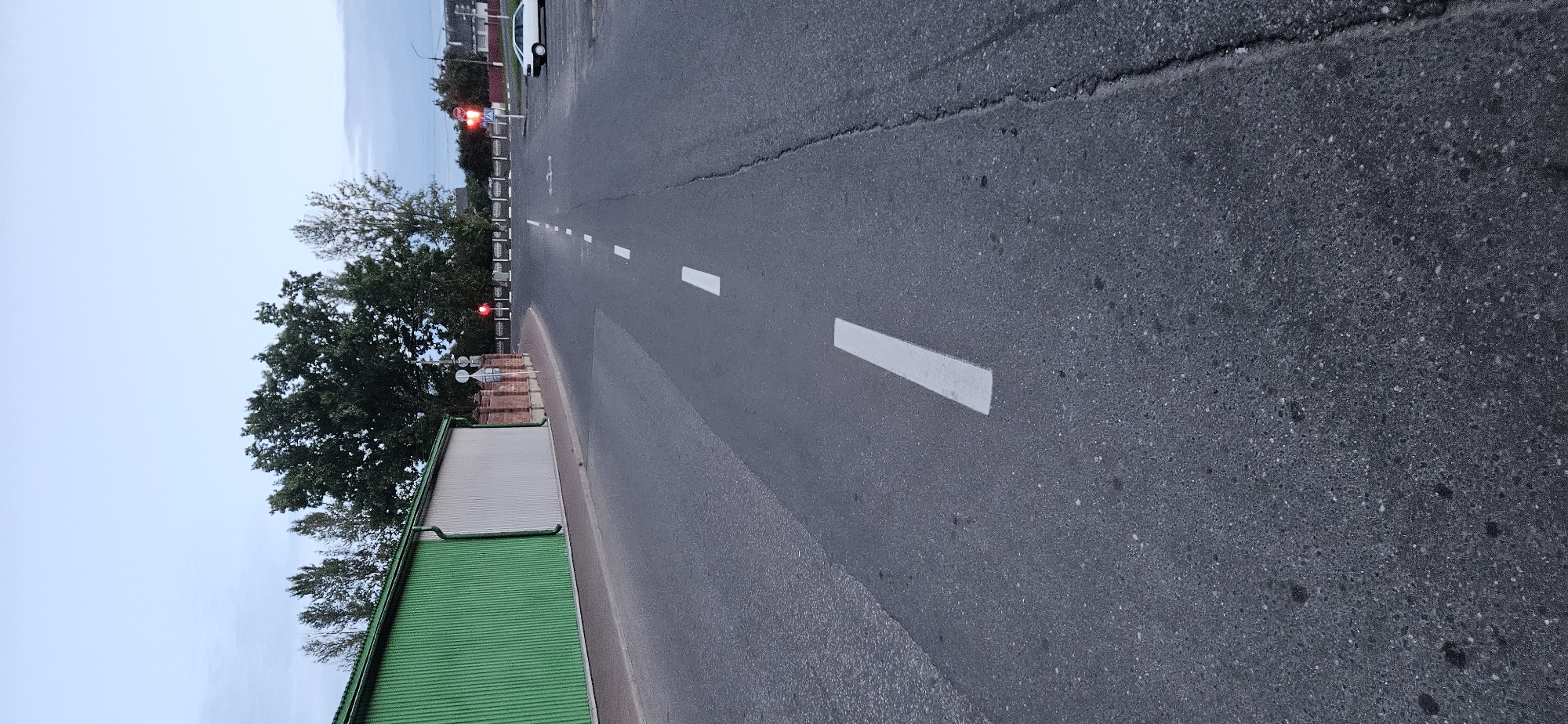 The image depicts an asphalt road with white dashed lane markings, leading towards an intersection. On the left side of the road is a long, corrugated green building adjacent to a lighter colored section. A brick wall separates the sidewalk from the building. Trees are visible behind the wall and building. In the distance, traffic lights show red, indicating a stop. A white car is parked on the right side of the road, near the intersection. The sky is overcast and blue. The overall scene suggests an outdoor urban environment, possibly in Vitsebsk, Belarus, during twilight.
