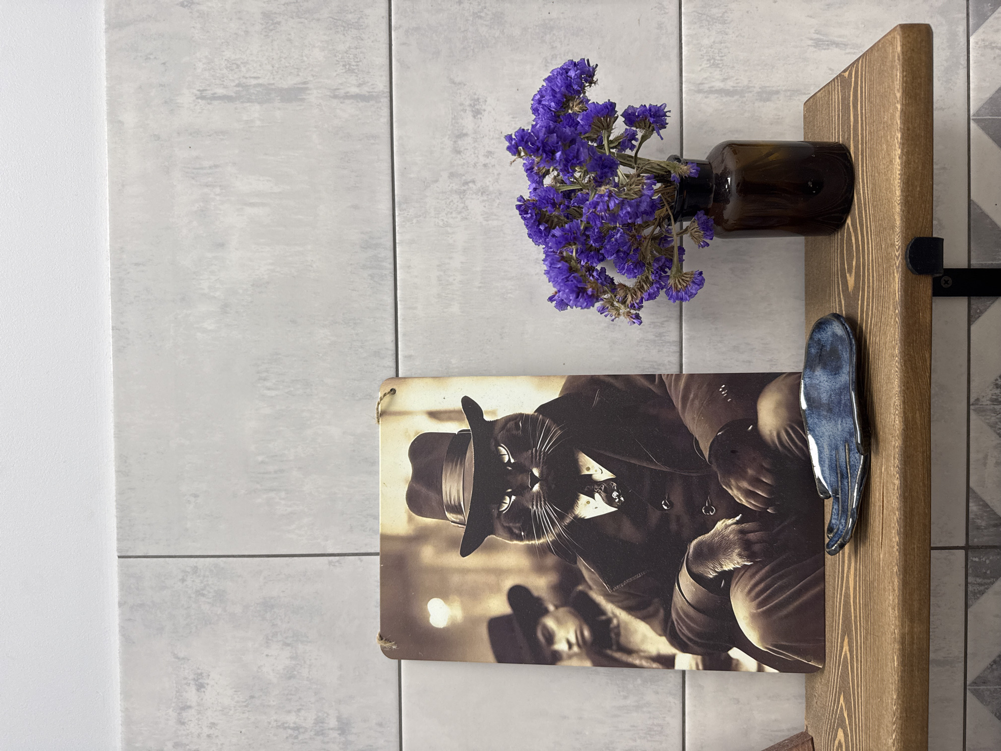 A sepia-toned photograph of a black cat wearing a fedora and a suit is displayed on a wall. The cat is looking directly at the viewer with a serious expression. Beside the photograph, a dark brown glass bottle with dried purple flowers is mounted on a wooden board. A dark blue ceramic hand-shaped object is also attached to the wooden board. The wall appears to be tiled with light gray and white tiles. The overall scene suggests a decorative arrangement, possibly in a home or a retail space. There is no visible text in the image.