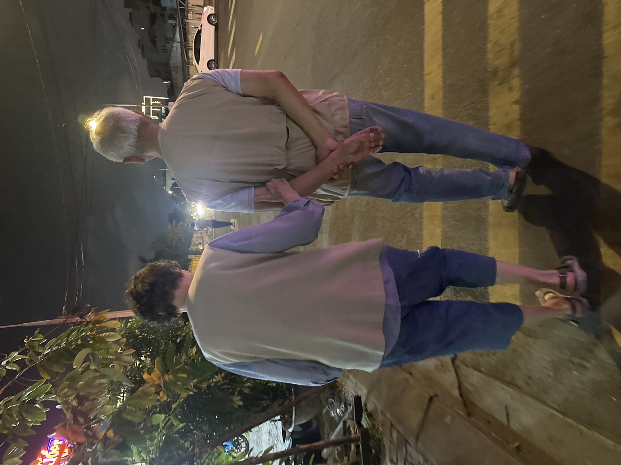 Two people are standing on a sidewalk in a brightly lit area of Đà Nẵng, Vietnam. Both are wearing casual clothing, including tan vests and blue shirts. One person is wearing jeans and dark shoes. The other is wearing blue cropped pants and sandals. Their arms are intertwined as if they are holding hands or embracing. The sidewalk has yellow stripes and is adjacent to a street with parked cars. There are trees and signage visible in the background, including a lit sign with what appear to be Vietnamese characters. The lighting is bright and there is a building visible in the background.