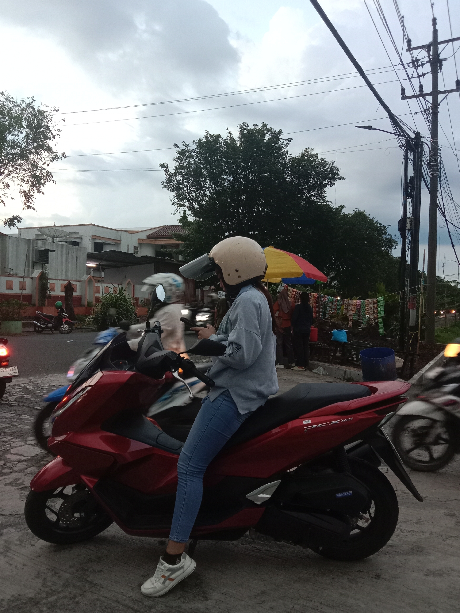 The image shows a woman on a red Honda PCX 160 scooter in Semarang, Indonesia. She is wearing a helmet, a light blue shirt, and blue jeans, and her right foot is resting on the ground. Other motorbikes are visible in front and to the right of her, and a building and trees are in the background. A yellow and red umbrella is set up over a roadside stall. A utility pole with wires is in the right background. The sky is overcast.