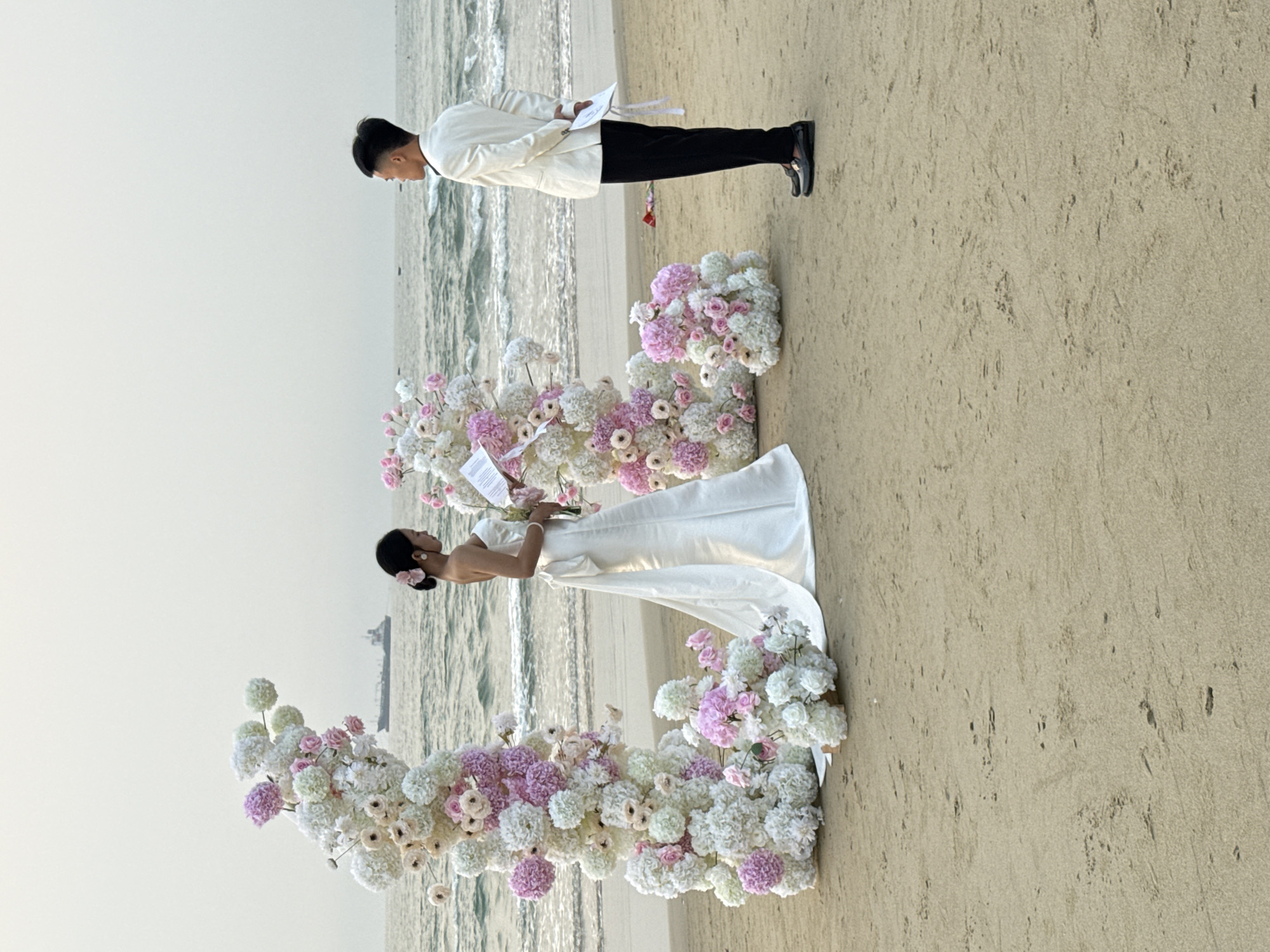 A man and woman are standing on a beach, seemingly during a wedding ceremony. The woman is wearing a white dress and the man is wearing a light-colored jacket with black pants. They are both holding papers, possibly vows. An elaborate floral arrangement of white and pink flowers is positioned in front of them, in a U shape. The beach setting includes sand and ocean waves. A ship can be seen in the background. The photo appears to be taken in Đà Nẵng, Vietnam based on the recognizable geographical elements.