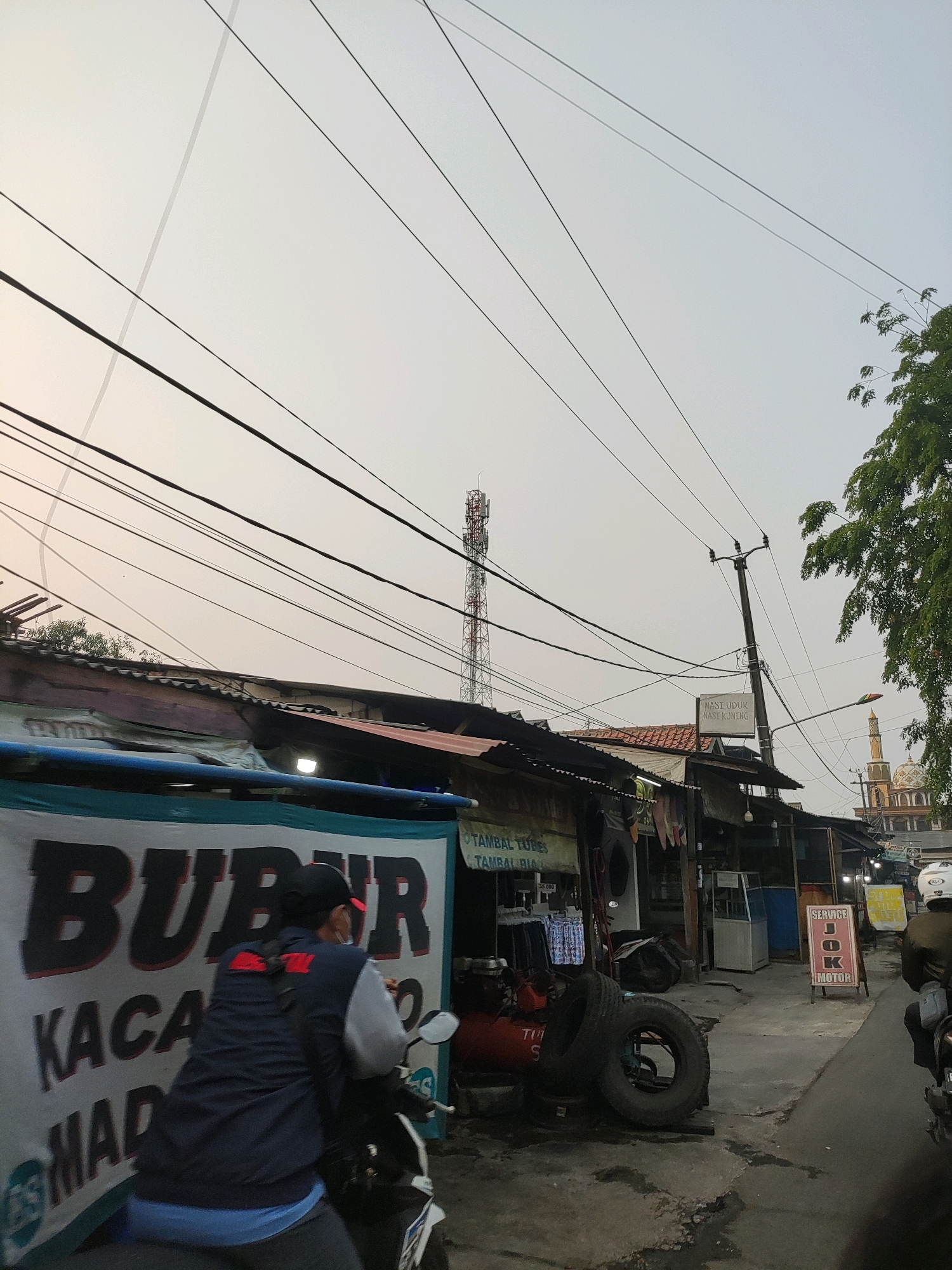 In Wanajaya, Indonesia, a street scene unfolds under an overcast sky, marked by numerous power lines crisscrossing overhead. A person on a motorcycle, wearing a dark blue vest over a light blue shirt and a black cap, passes in front of a large banner advertising "BUBUR KACANG MADU". The banner is partially obscured by the motorcycle and rider. To the right of the banner, several small shops line the street. One shop has a sign reading "TAMBAL TUBES TAMBAL RUSA". Further down the street, another shop displays a sign that says "NASI UDUK NASI KUNING". A cell tower stands prominently in the background, and a mosque with a dome and minaret is visible in the distance on the right. A red air compressor with tires is placed outside one of the shops. Another motorcycle with a rider wearing a white helmet is seen further down the street. The overall environment suggests a busy, working-class commercial area.