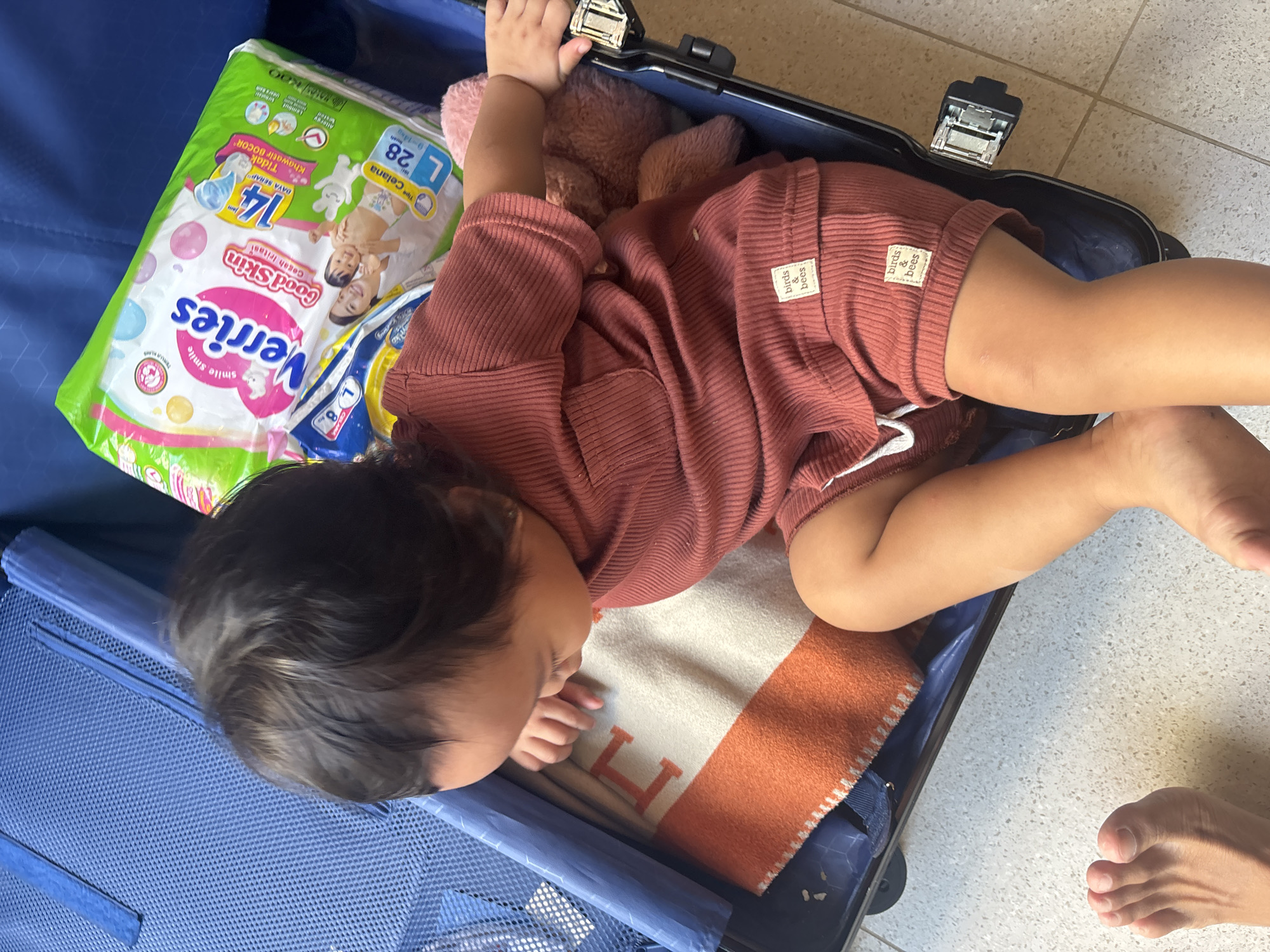 A toddler with dark hair, wearing a rust-colored ribbed knit outfit, is asleep in an open blue suitcase. A plush pink toy is visible behind the toddler. Next to the toddler, a package of "Verries" diapers and a white blanket with an orange border and a large orange "H" are present. The suitcase is on a tiled floor.