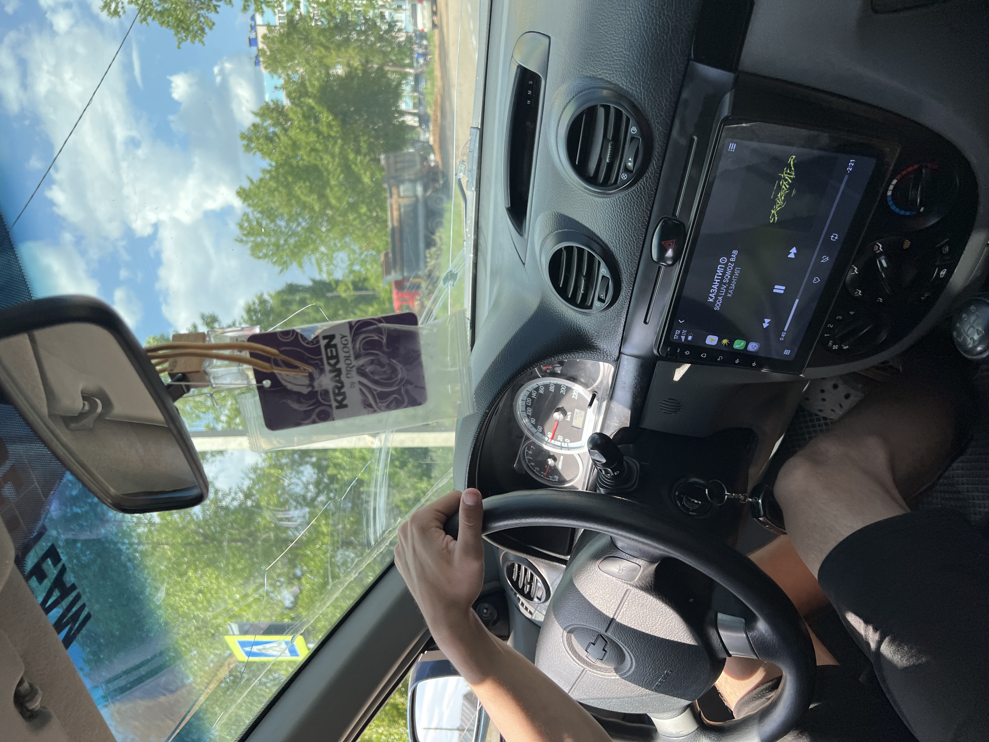 The image, taken from inside a car, shows the driver's perspective while holding the steering wheel. The car is a Chevrolet, evident from the logo on the steering wheel. The infotainment system is playing a track titled "КАЗАНТИП" by SODA LUV and SQWOZ BAB. The speedometer indicates the car is not in motion. An air freshener from Kraken by Prology hangs from the rearview mirror. A sticker is on the windshield. The windshield appears to be cracked. The car is in Chaikovsky, Russia. Outside, greenery and a truck are visible through the windshield. The sky is partly cloudy.