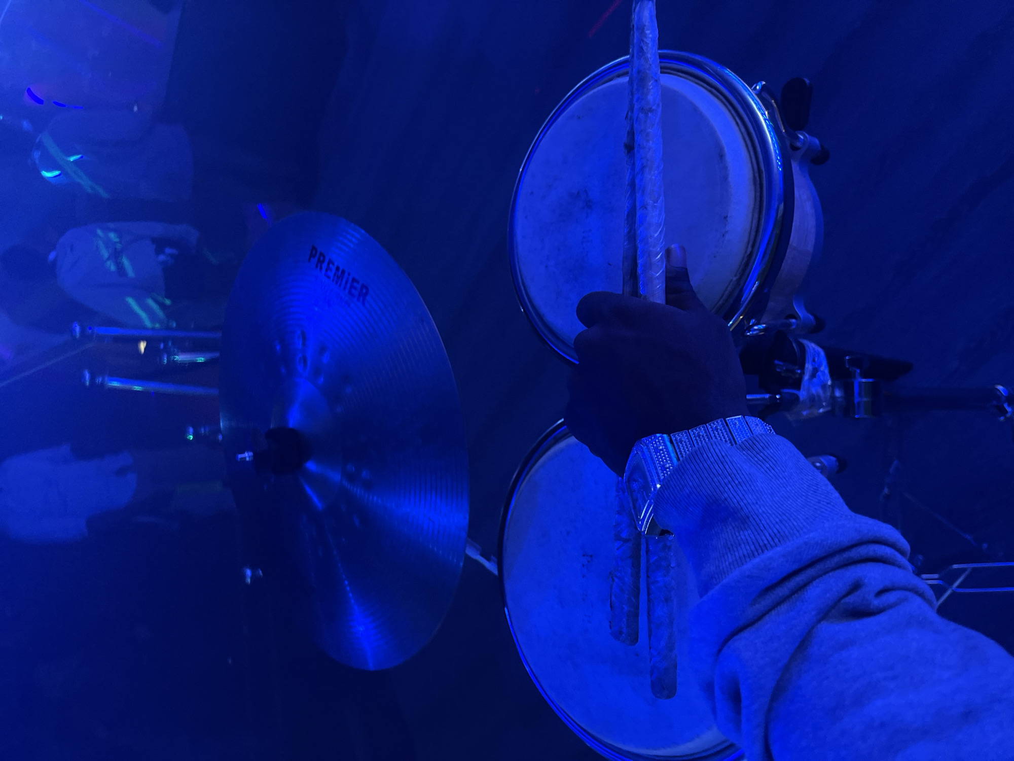 The image shows a person playing a set of drums, comprising a pair of bongo drums and a cymbal, likely in a dark room with blue lighting. The cymbal is labeled "Premier". The location is not discernible as Jalingo, Nigeria from the given visual information.
