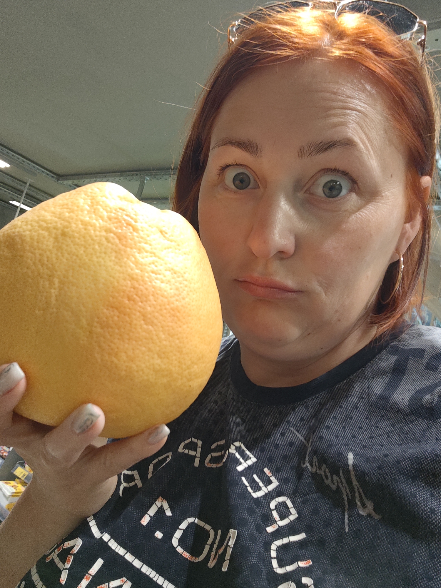 A woman with red hair and sunglasses on her head holds a large grapefruit close to her face. She has light skin, blue eyes, and wears gold hoop earrings. Her expression is one of surprise or skepticism. She is wearing a dark blue t-shirt with white lettering. The background appears to be an indoor commercial setting, possibly a grocery store, with shelving and ceiling fixtures visible. The photo appears to have been taken in the city of Марковский, Russia.