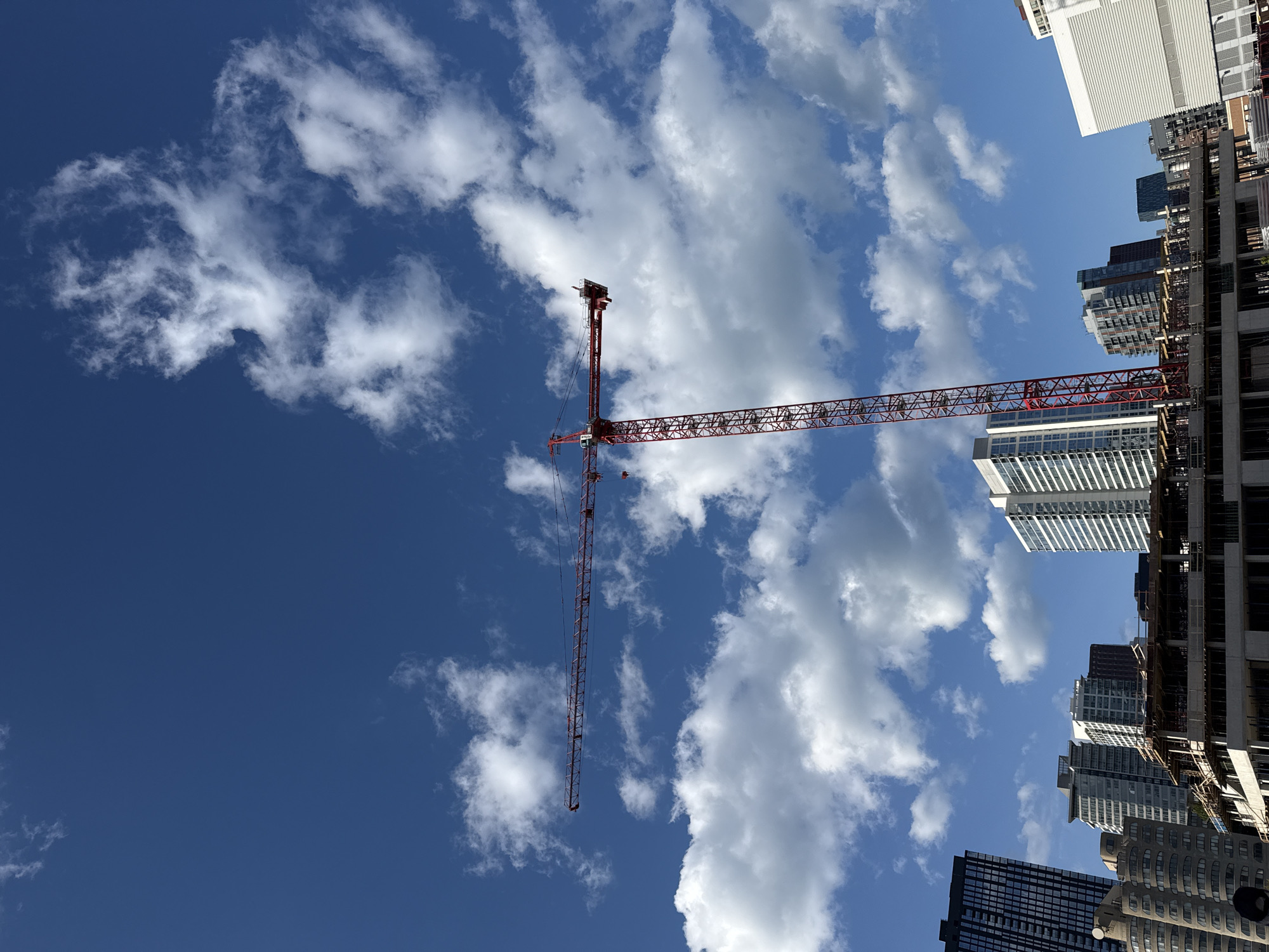 The image depicts a construction site in Chicago, USA. A red crane dominates the frame, stretching across a blue sky filled with scattered white clouds. The crane is positioned amongst several tall buildings, some of which are under construction. The scene appears to be taken from a low angle, looking upwards towards the sky and the towering structures.