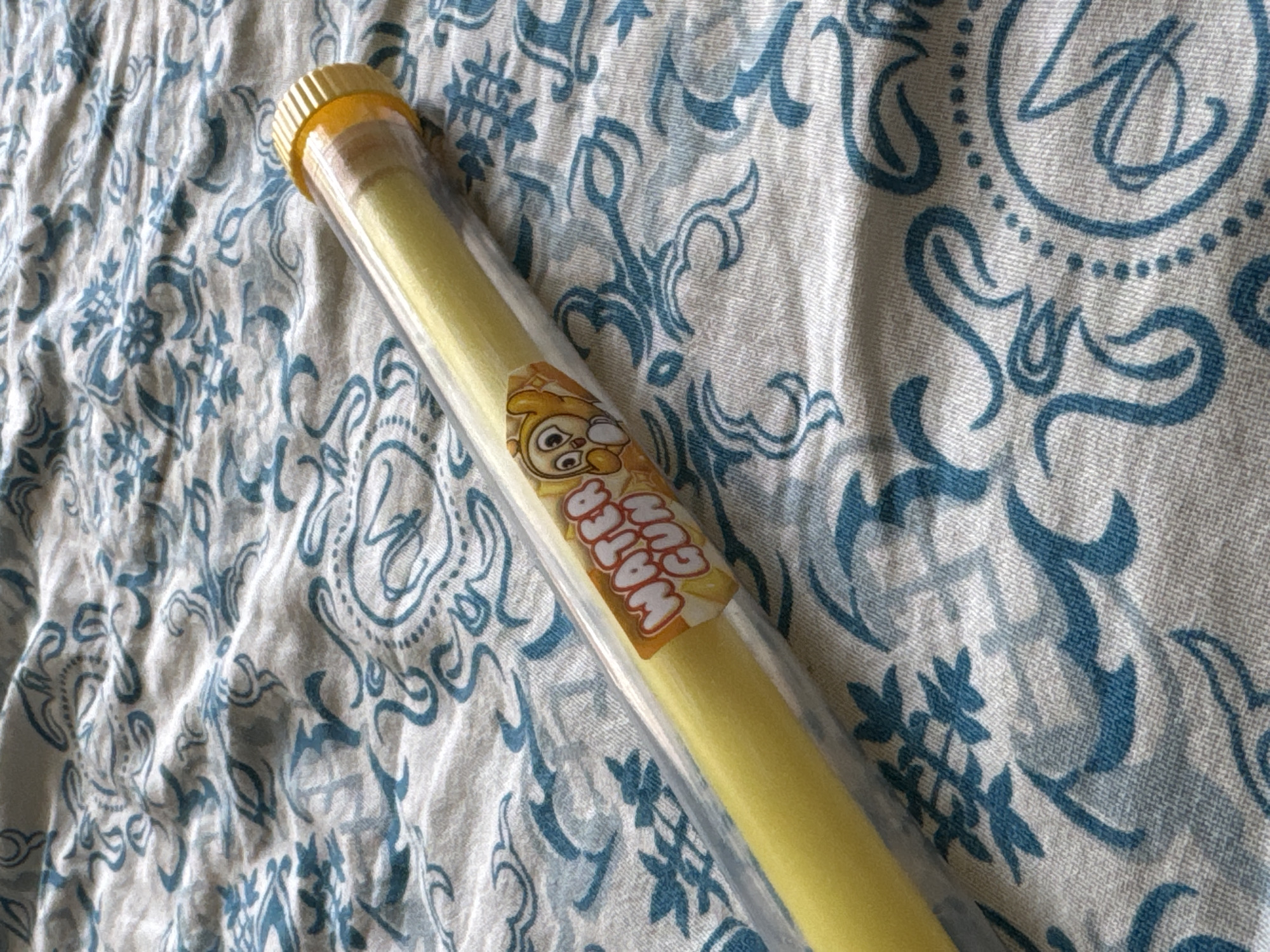A yellow water gun is enclosed in a clear plastic tube, resting on a fabric surface with a blue paisley pattern. The tube has a yellow cap. A sticker on the tube depicts a cartoon character and reads "WATER GUN". The fabric is wrinkled, suggesting it may be a blanket or bedspread. There are no people present in the image. The scene is indoors and appears to be a still life.