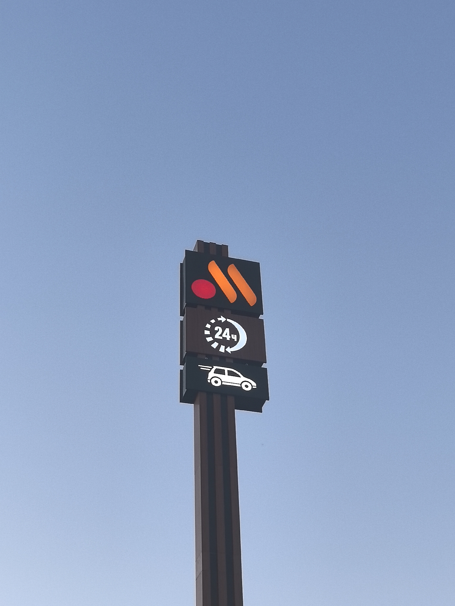 A tall, dark brown signpost stands against a clear blue sky. The signpost has multiple rectangular panels attached to its upper section. The topmost panel displays an orange "M" logo with a red circle to its left. Below this, a panel shows a white circular icon with the number "24" in the center, enclosed by arrows, indicating 24-hour service. The lowest visible panel features a white silhouette of a car. The signpost is located on the side of a road, with hints of dark brown vertical structures suggesting a structure or building behind it. The overall scene is set in an outdoor urban environment, possibly in the city of Krasnodar, Russia, given the context of the branding displayed. There are no people visible in the image.