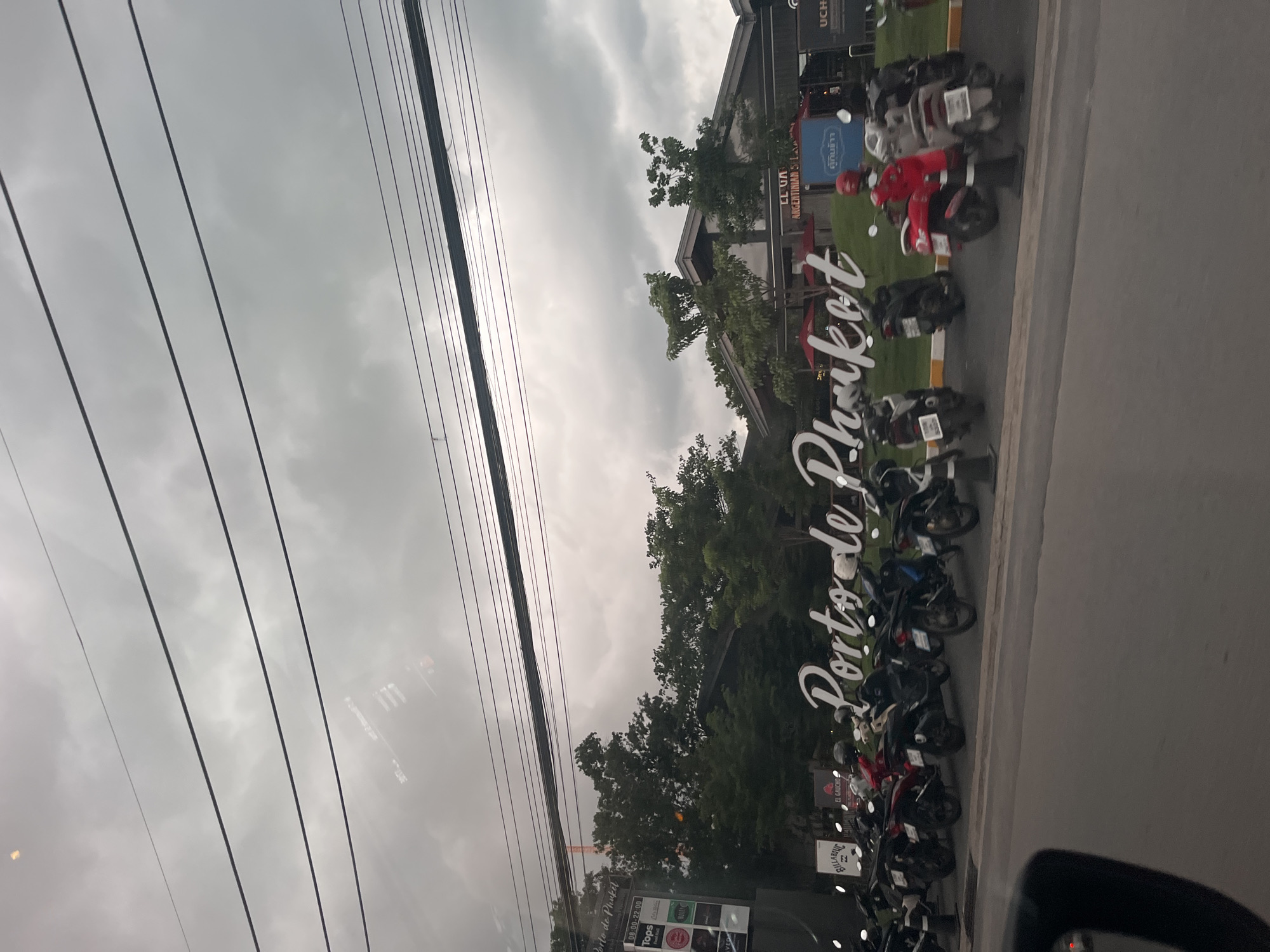 The image shows a cloudy sky over a street scene in Choeng Thale, Thailand. In the foreground, a row of parked motorcycles is visible. Behind the motorcycles, a large, white sign reads "Porto de Phuket". Further in the background, buildings with sloped roofs are surrounded by trees. A smaller sign on the left side of the image contains various logos, including "Tops". The overall atmosphere is overcast and appears to be late afternoon or evening.
