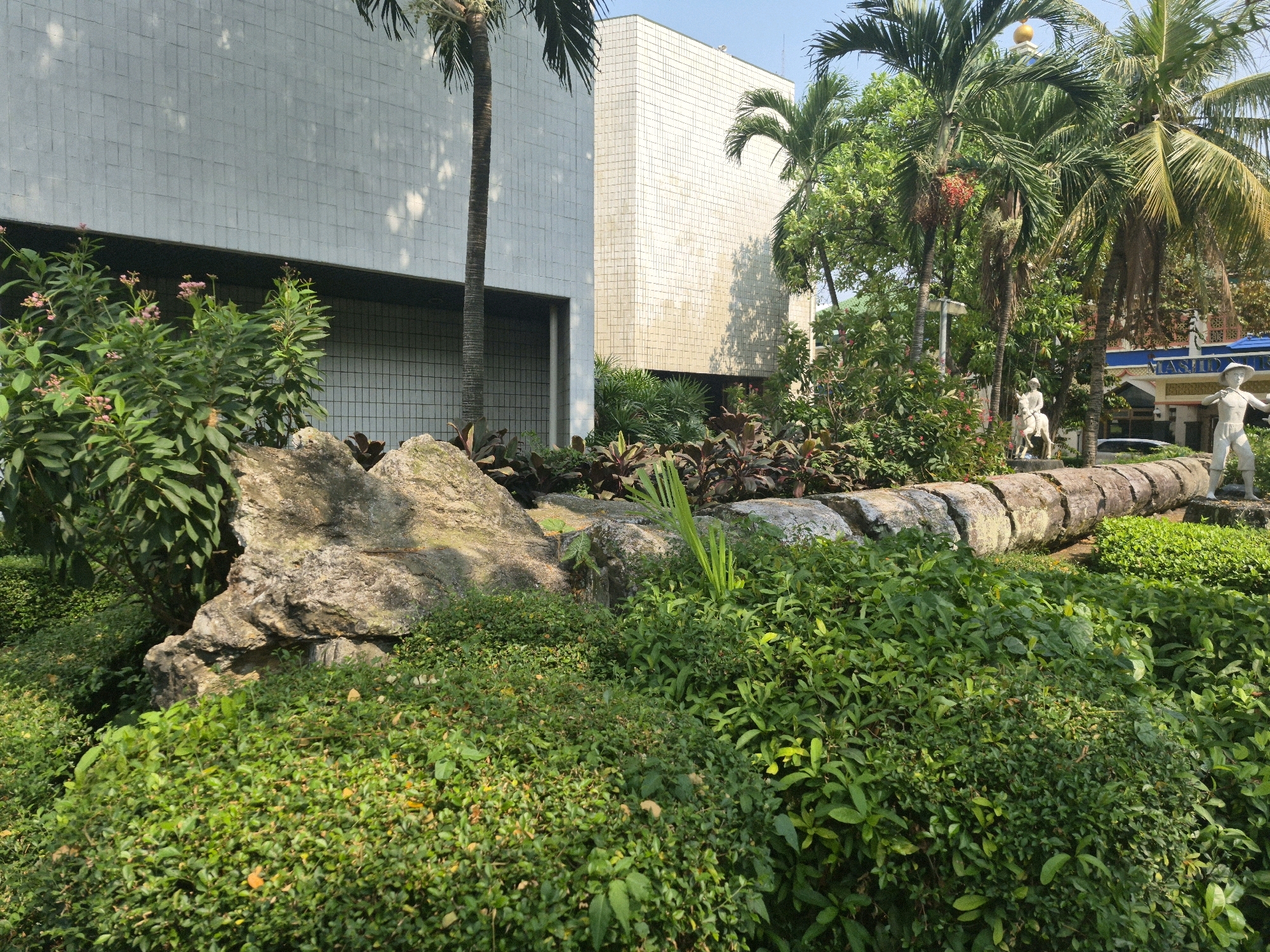 The image depicts the exterior of the National Museum of Indonesia in Central Jakarta, Indonesia. The museum's white, tiled facade is visible, with a tall palm tree in the foreground. A garden with lush green foliage and rocks is in front of the building. A large, segmented concrete structure resembling a fallen pillar lies horizontally across the garden. Two stone statues, one of a person on horseback and another of a standing person, are present in the garden. The scene is bathed in sunlight, creating shadows on the building and foliage.