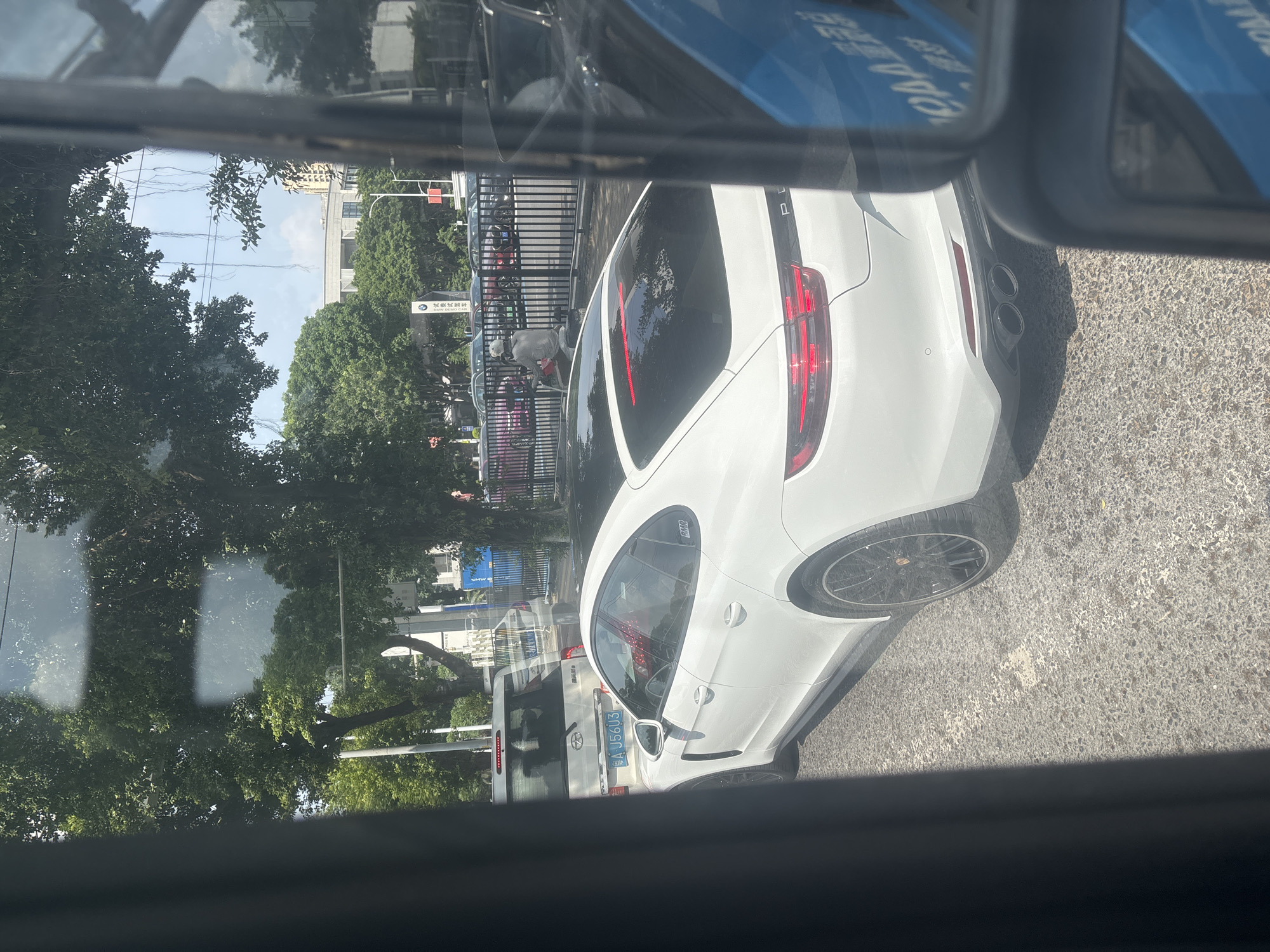 A white Porsche Panamera is parked on a city street in Baiyun District, China. The car is facing away from the camera, with its rear end visible. A white SUV with a blue license plate is parked behind the Porsche. In the background, there are trees, buildings, and a black metal fence. A person on a motorcycle is visible in front of the fence. The scene appears to be a sunny day, with clear skies and shadows cast by the trees. The image is taken from inside another vehicle, as evidenced by the reflection in the side mirror and the framing of the shot. The license plate on the white SUV reads "A J56U3". The Porsche has a black roof and black rims. The dual exhaust pipes of the Porsche are visible.