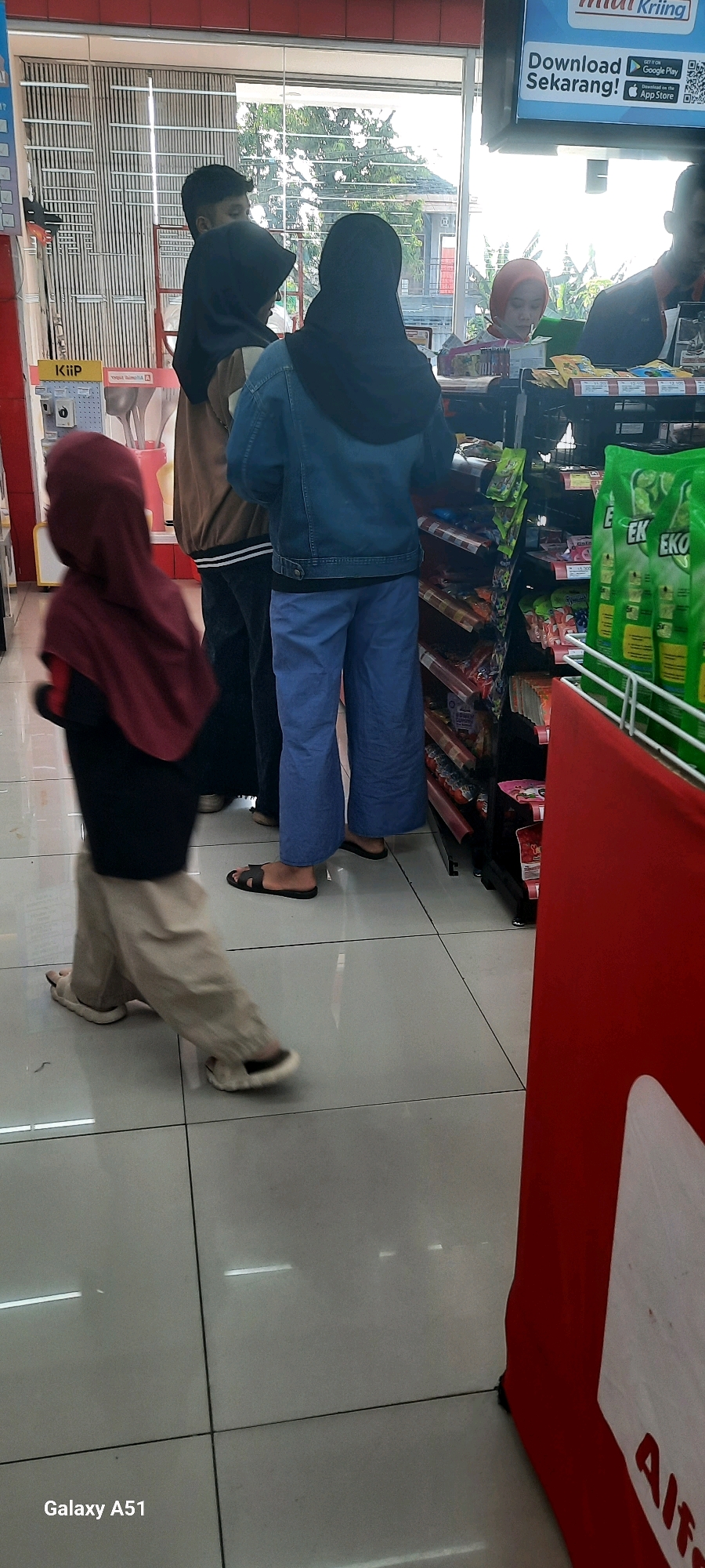 The image shows a street scene in Wlingi, Indonesia, likely outside a convenience store. In the foreground, a child in a maroon hijab and beige pants walks on a tiled floor. Behind the child, two people, one in a dark hijab and denim jacket, and another in a tan jacket, are standing. Further back, a person in a red hijab is at a counter, possibly a cashier. Shelves stocked with various products, including what appear to be cleaning supplies in green packaging, are visible in the background. The environment suggests a commercial area with bright lighting and retail displays.