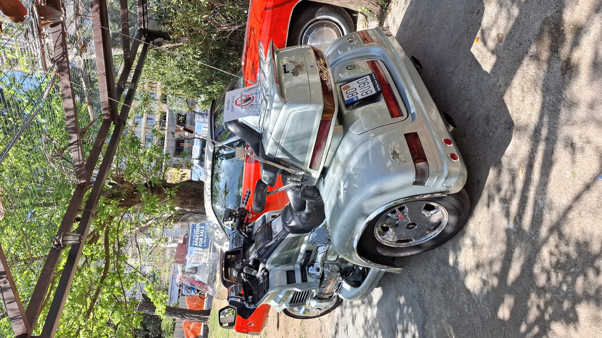 The image shows a pale blue Gold Wing motorcycle parked on a concrete surface next to a red car. A sign on the motorcycle reads "NE DIRAJ / DON'T TOUCH" and displays a license plate with "BD BU900". In the background, foliage and a metal structure are visible, along with a sign advertising "APARTMENTS FOR SALE."